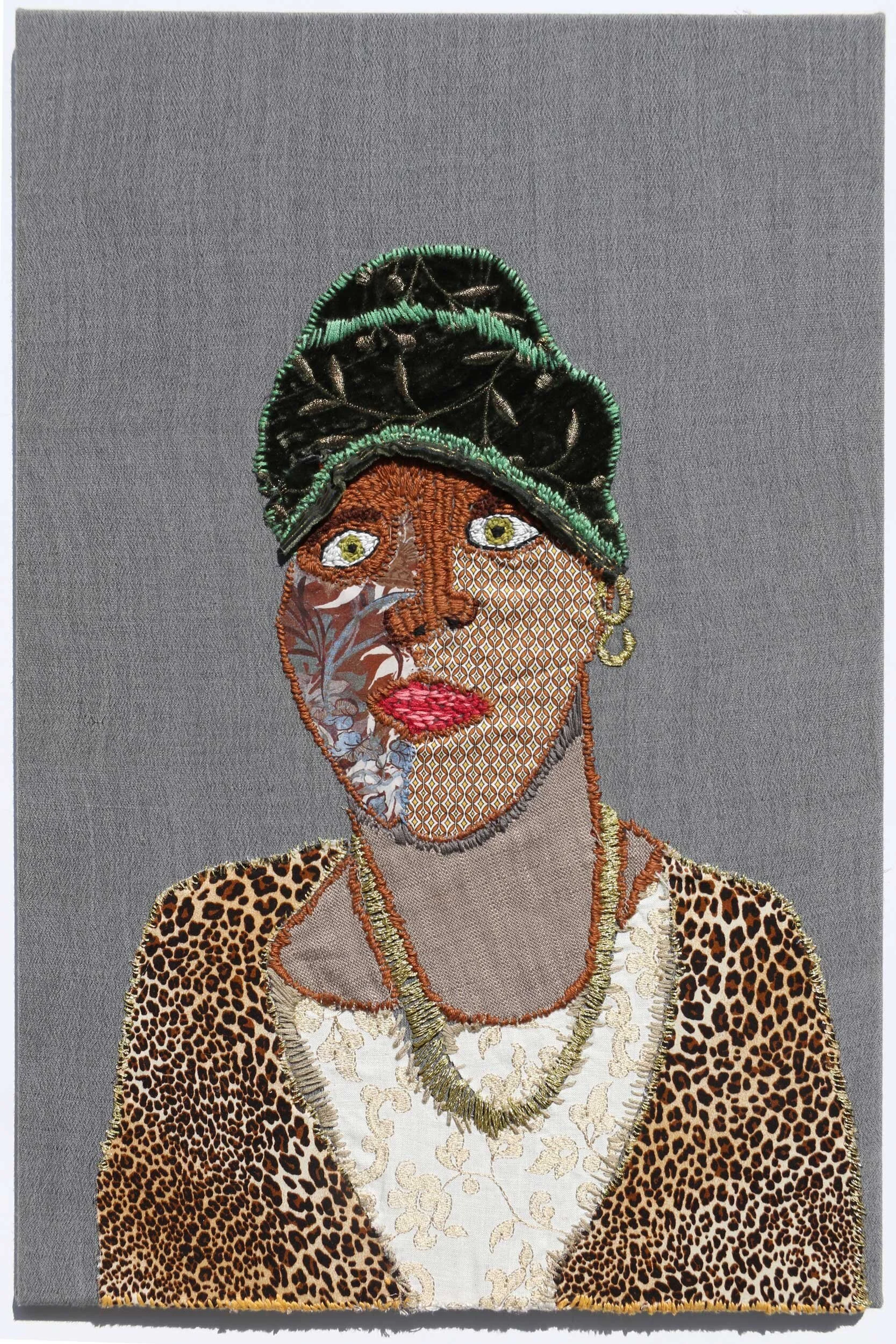 A grey cloth backdrop with a figure from waist up featured. The lady has brown skin made from different fabrics, a leopard print cardigan and a green/black hat on. All embroidered on top.