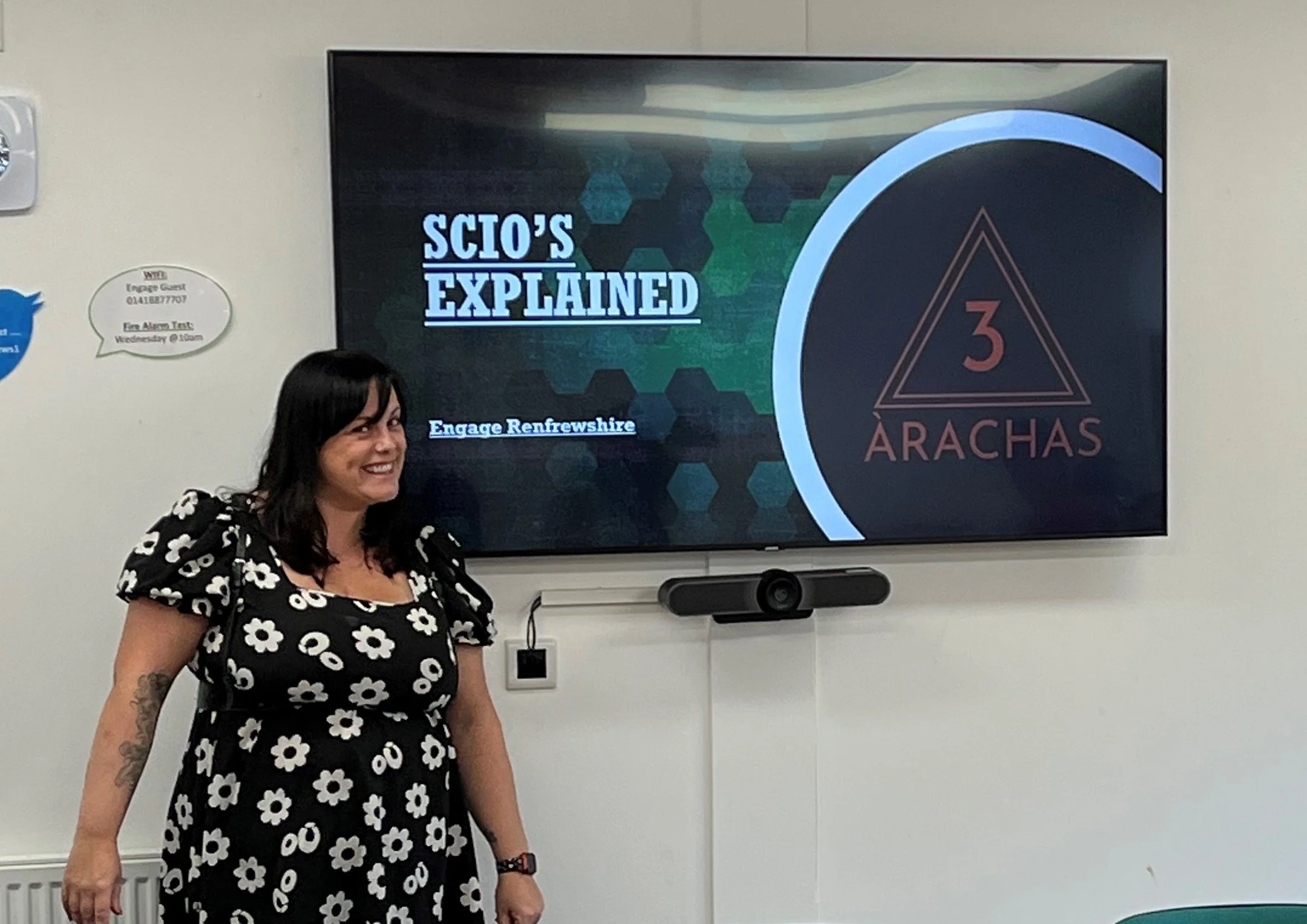 A woman standing in front of a screen with the title "SCIO'S EXPLAINED" and a logo with a triangle and the number 3 with the text "ARACHAS" on it, in a meeting room.