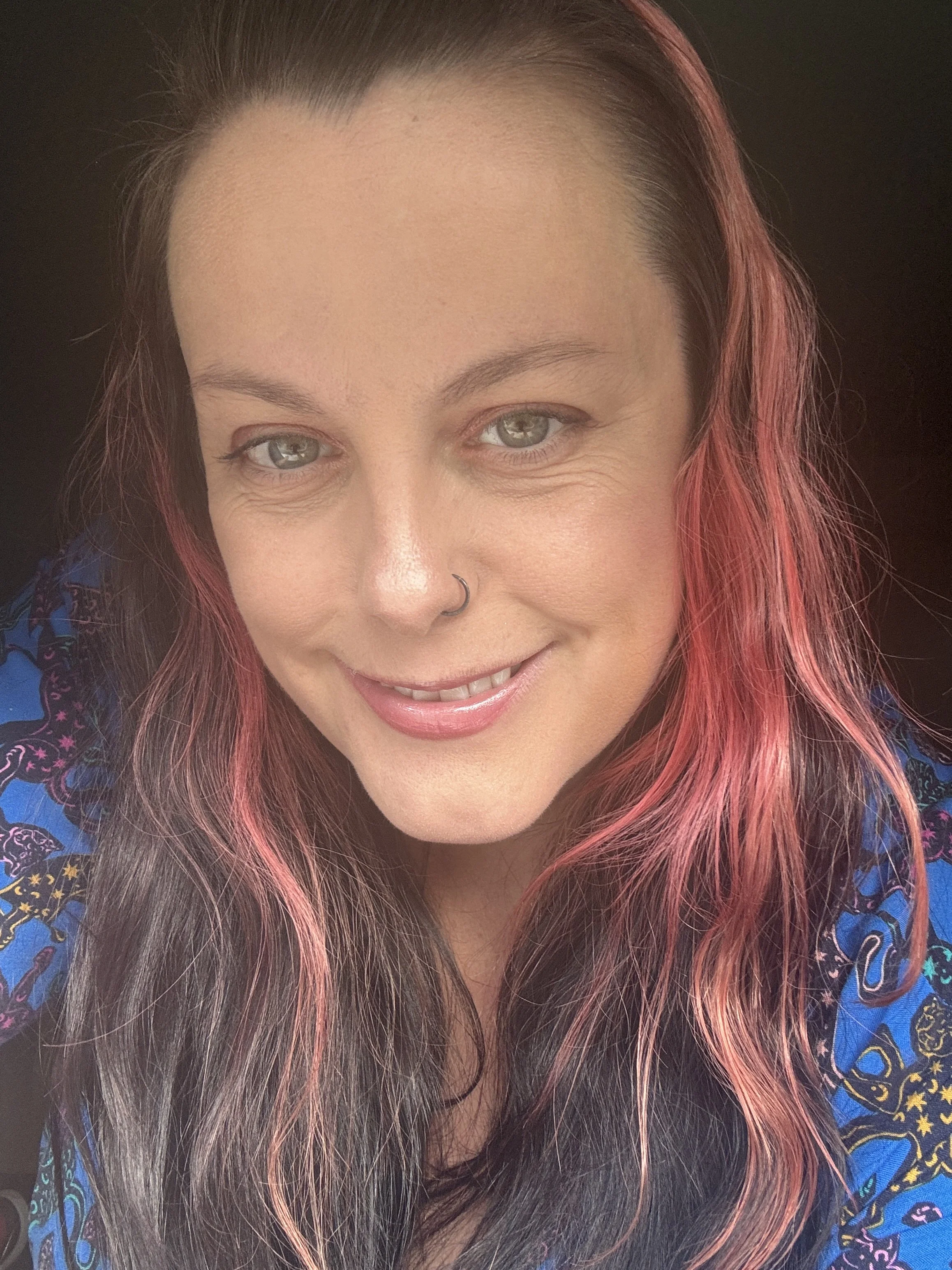 Close-up of a smiling woman with green eyes, pink and black hair, wearing a blue patterned top, and a silver nose ring.