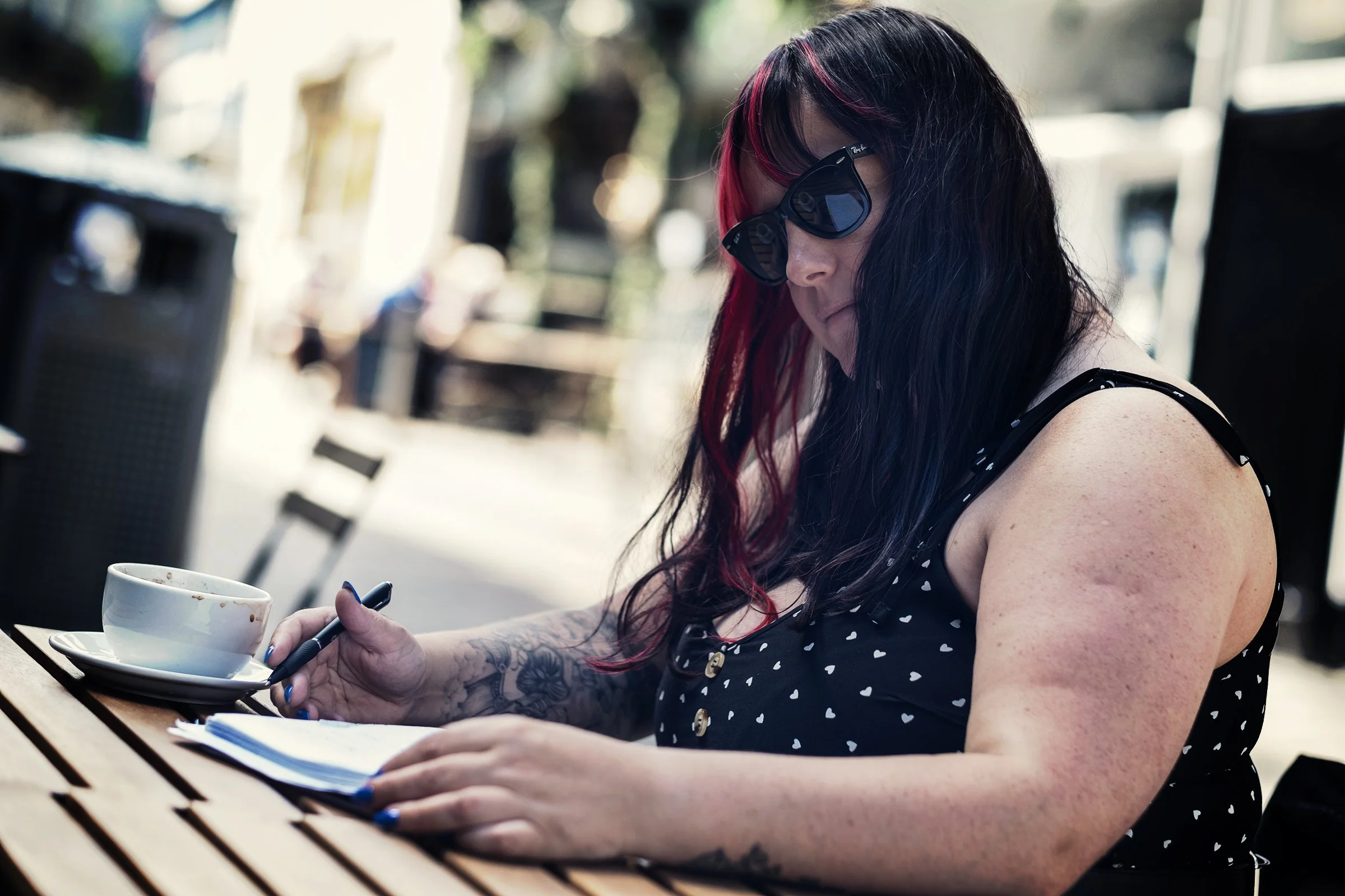 A woman with dark hair and red streaks wearing sunglasses, a black sleeveless dress with white hearts, is sitting outdoors at a wooden table, writing in a notebook with a pen, with a cup of coffee nearby.