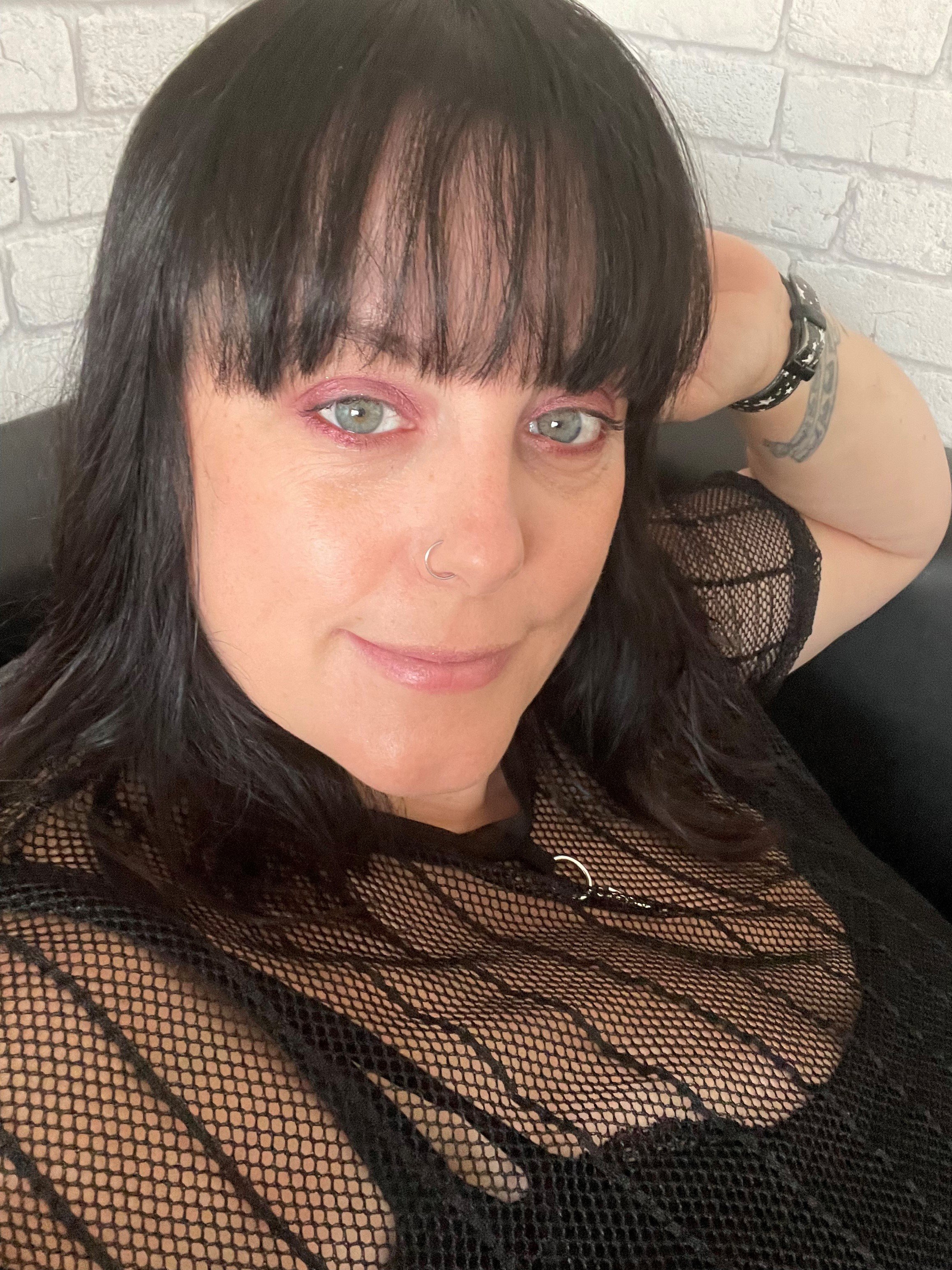 A woman with black hair and bangs, light blue eyes, wearing a black fishnet top, a nose ring, and a black wristwatch, sitting on a black sofa against a white brick wall, smiling softly.