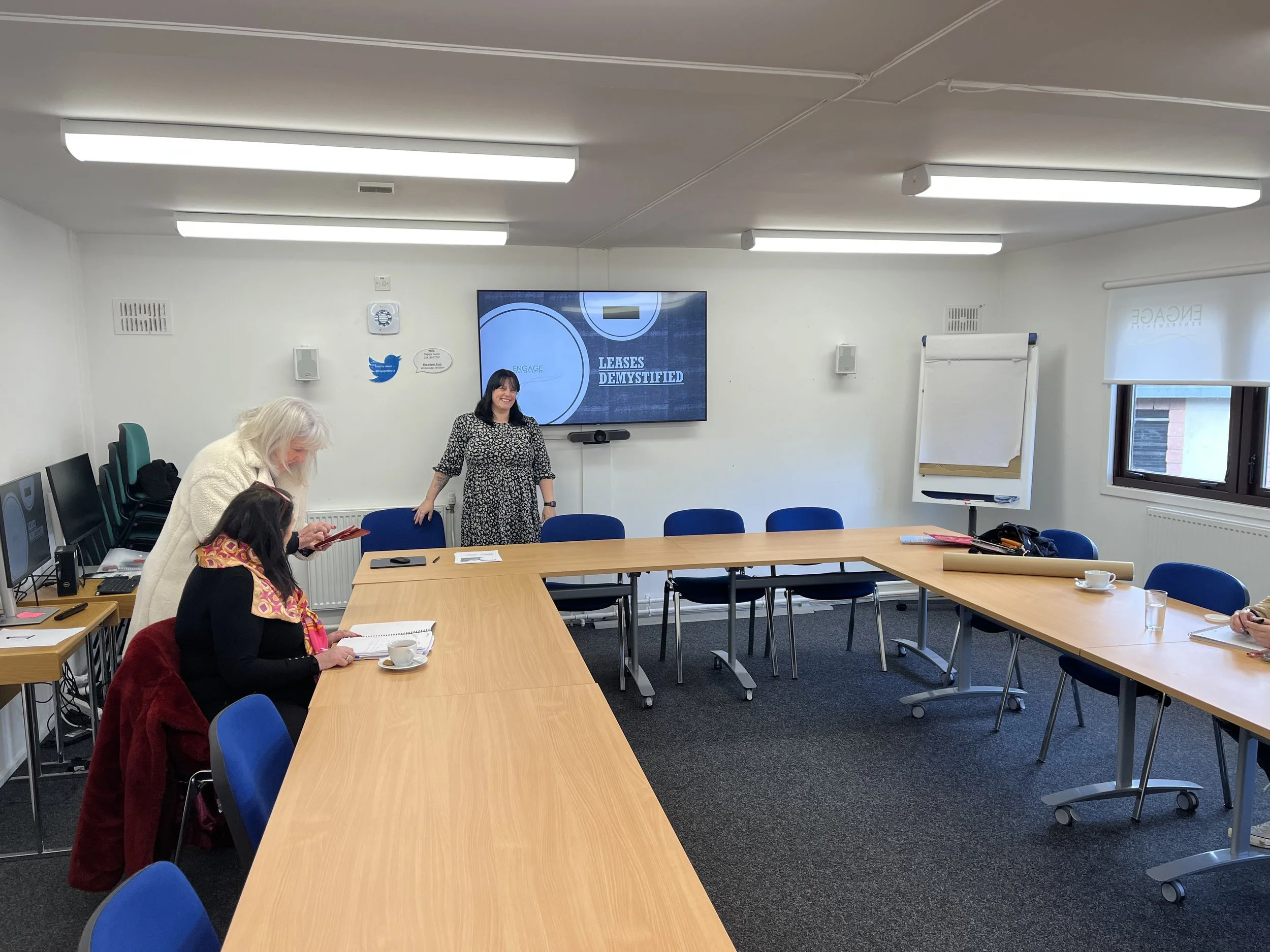 A meeting room with four women preparing for a presentation. One woman stands near a large screen that reads 'LEASES DEMYSTIFIED', while others are seated at tables with notebooks and cups.