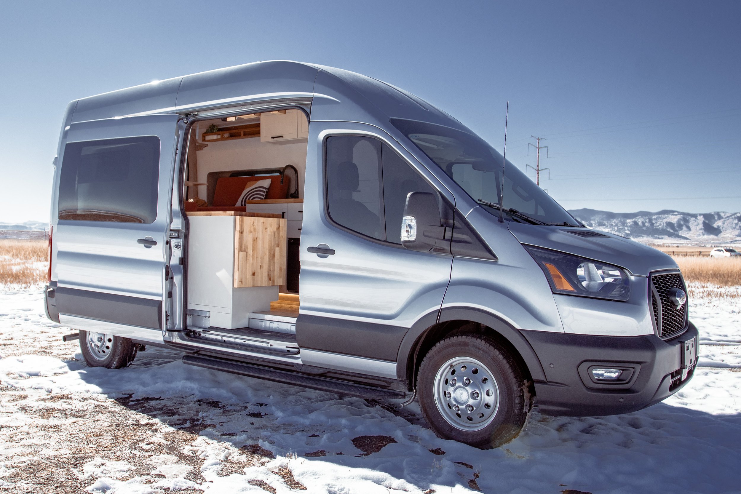 Silver camper van with open sliding door showing interior on snowy ground with mountains in background.