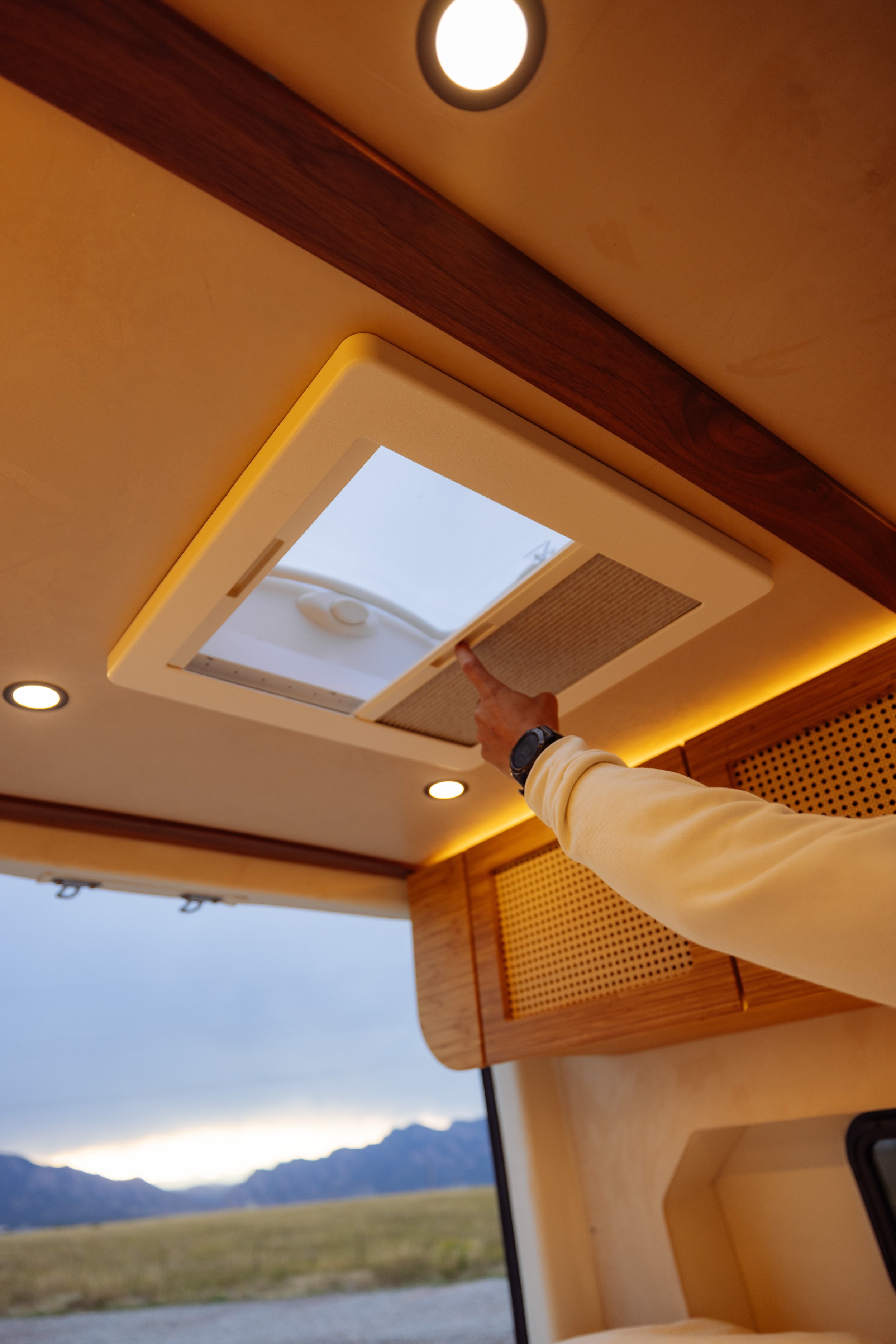 Person's hand adjusting a ceiling skylight in a van interior with wooden accents and landscape view outside.
