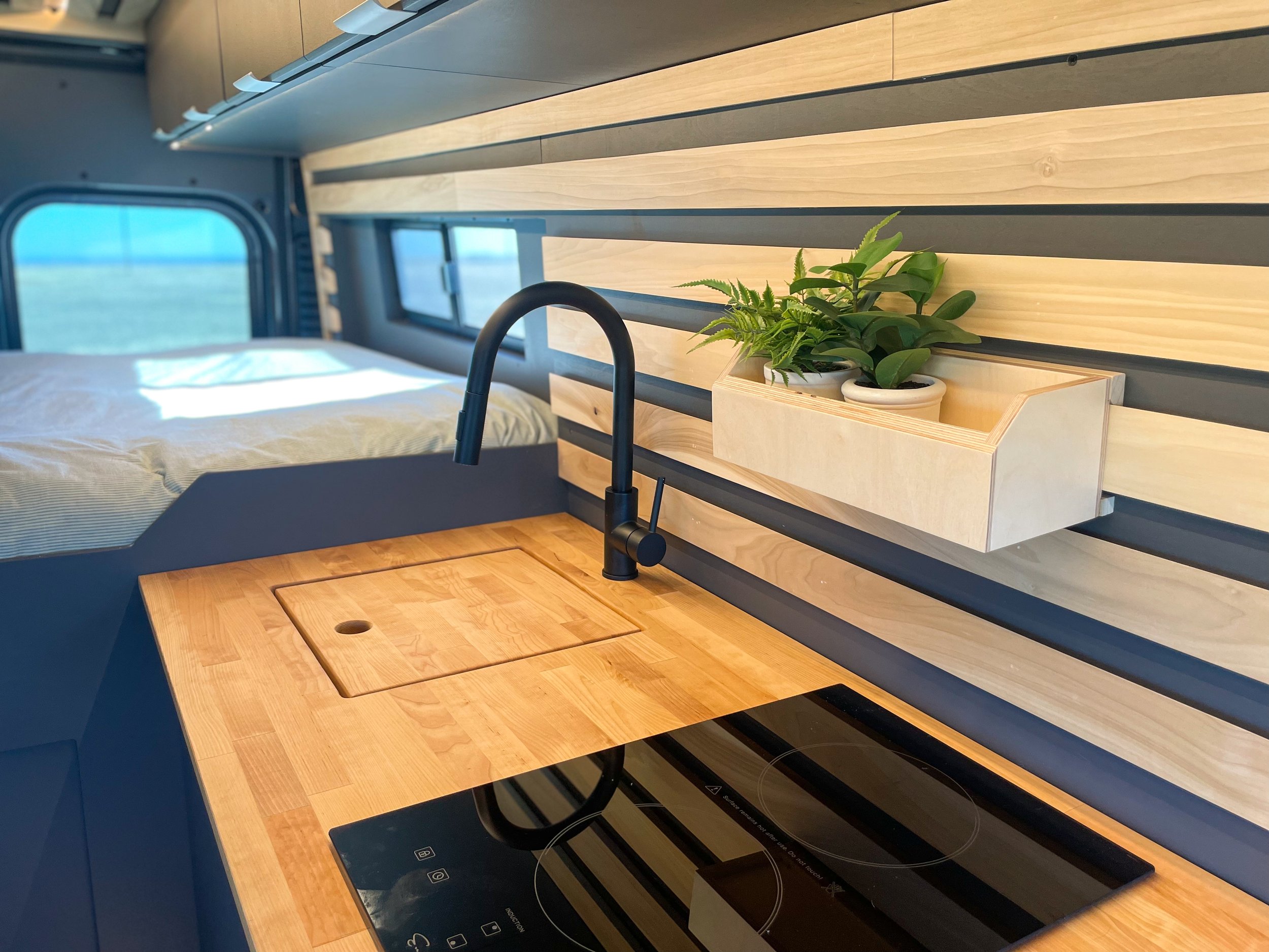 Interior of a modern camper van with a wooden countertop and induction cooktop, a black faucet, and a wooden planter box with small plants. The area features striped wooden walls and a bed in the background.