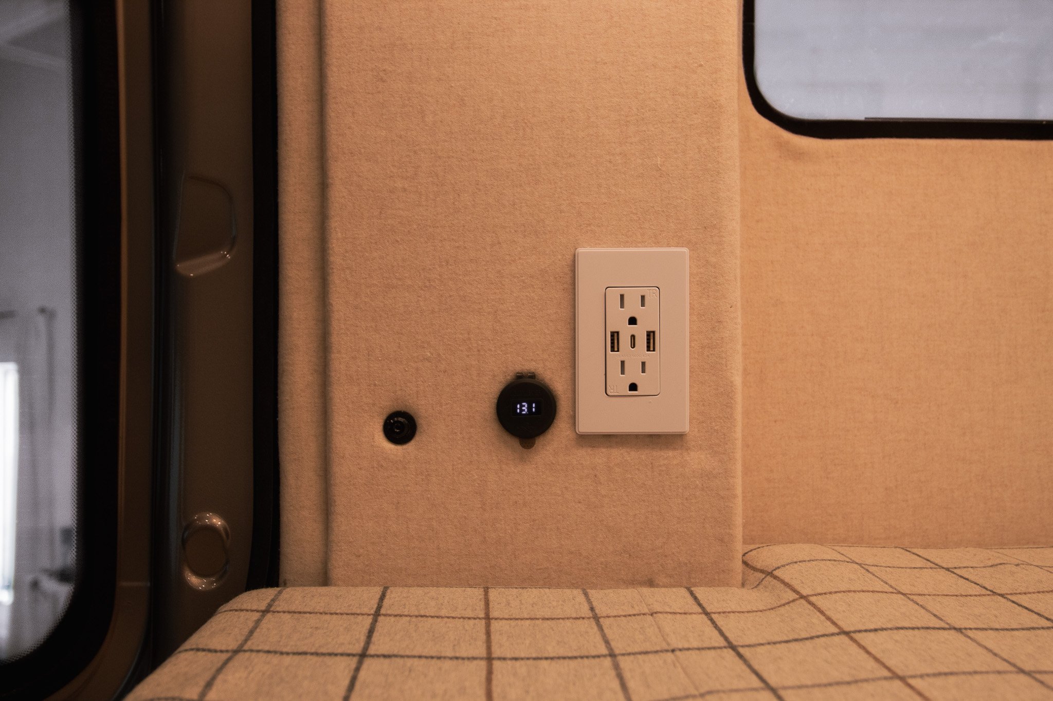 Interior of a vehicle with beige fabric walls, featuring a standard electrical outlet with USB ports, black round gauge displaying "13.1", and a window on the left.