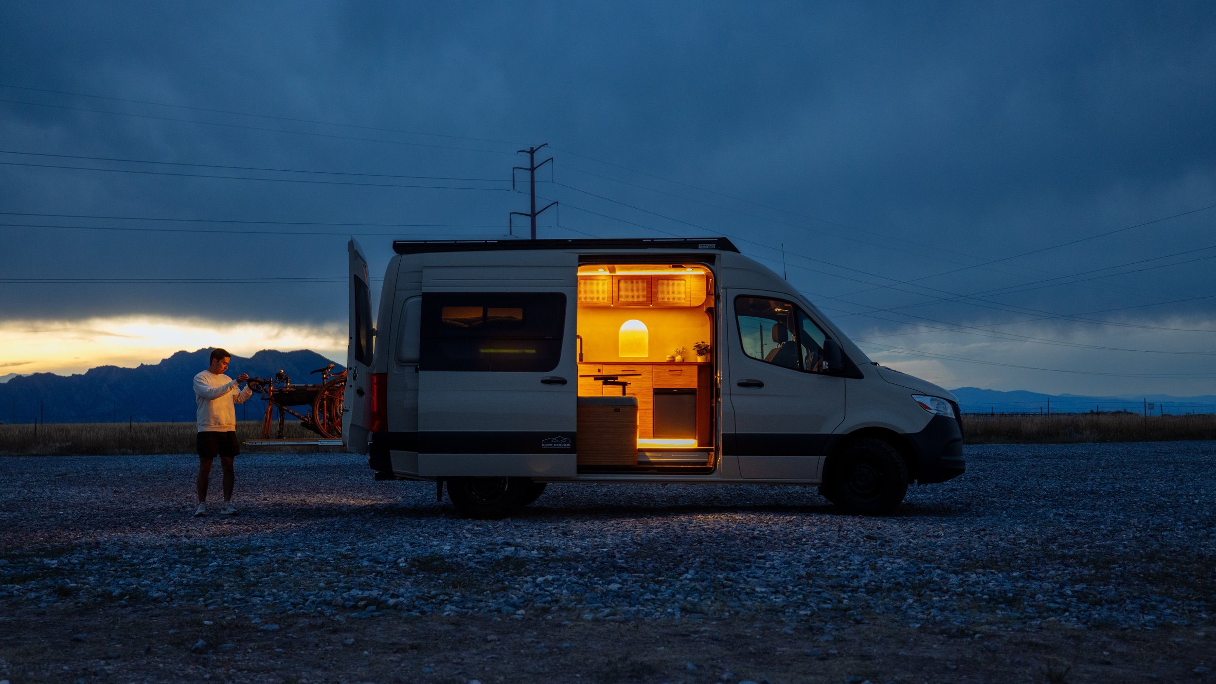 A camper van with its side door open, revealing a lit interior, parked in an outdoor setting at dusk. A person is standing near the back of the van, adjusting a bicycle mounted on the rear.