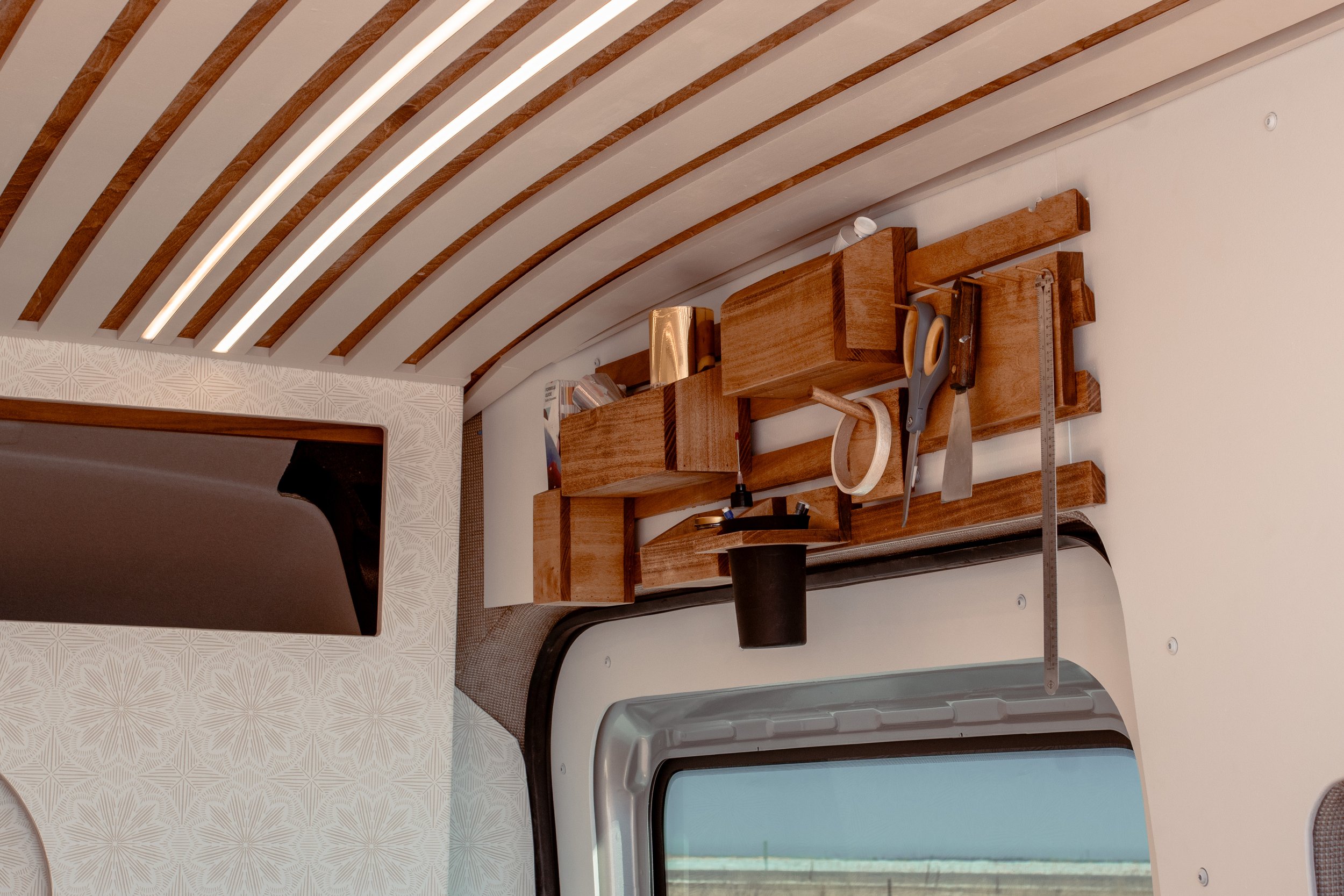 Interior of a van with wooden paneling, a wooden storage shelf holding various items like scissors and tape, and a window partially visible.