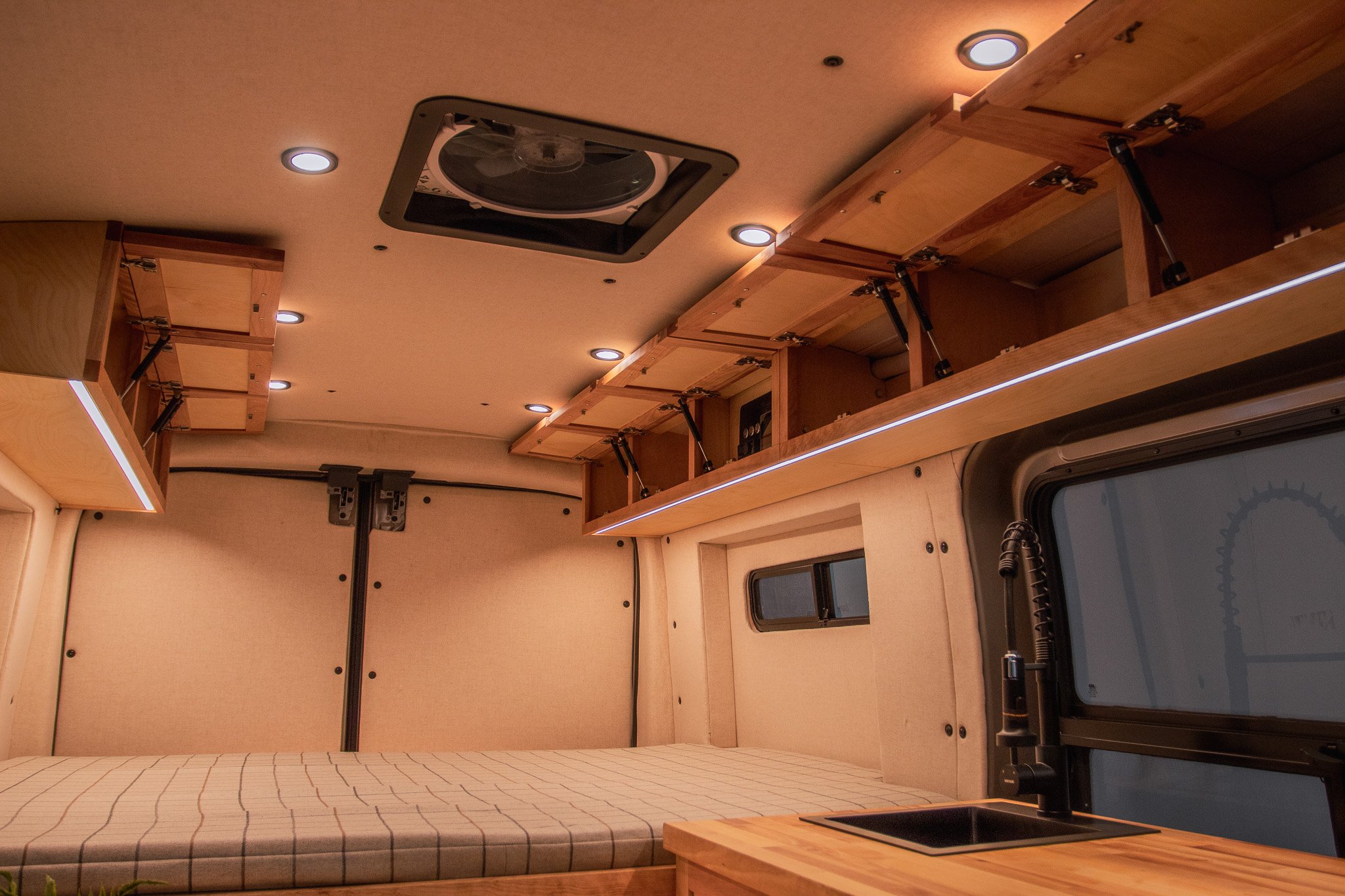 Interior of a camper van with wooden cabinets, a bed, overhead lighting, and a small sink.