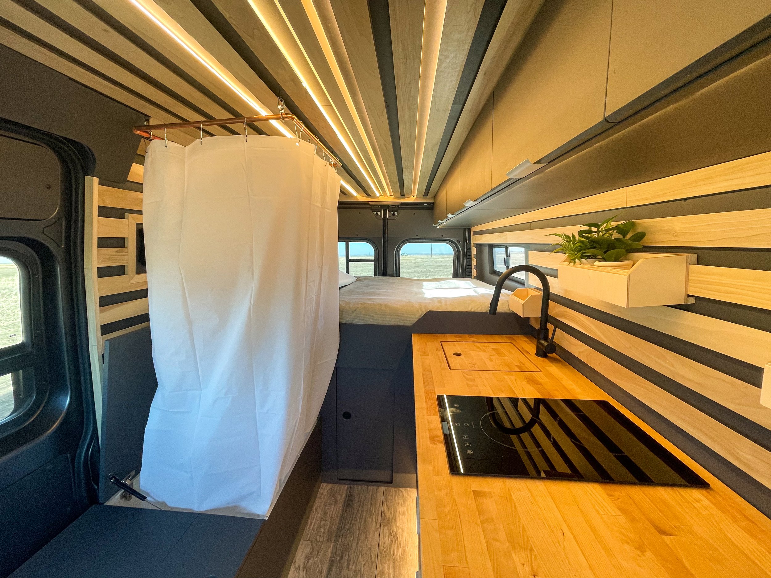 Interior of a modern camper van with a kitchenette featuring a wooden countertop and induction stove, a small sink, a white shower curtain, and a bed in the background. Wood paneling and a potted plant add decor.