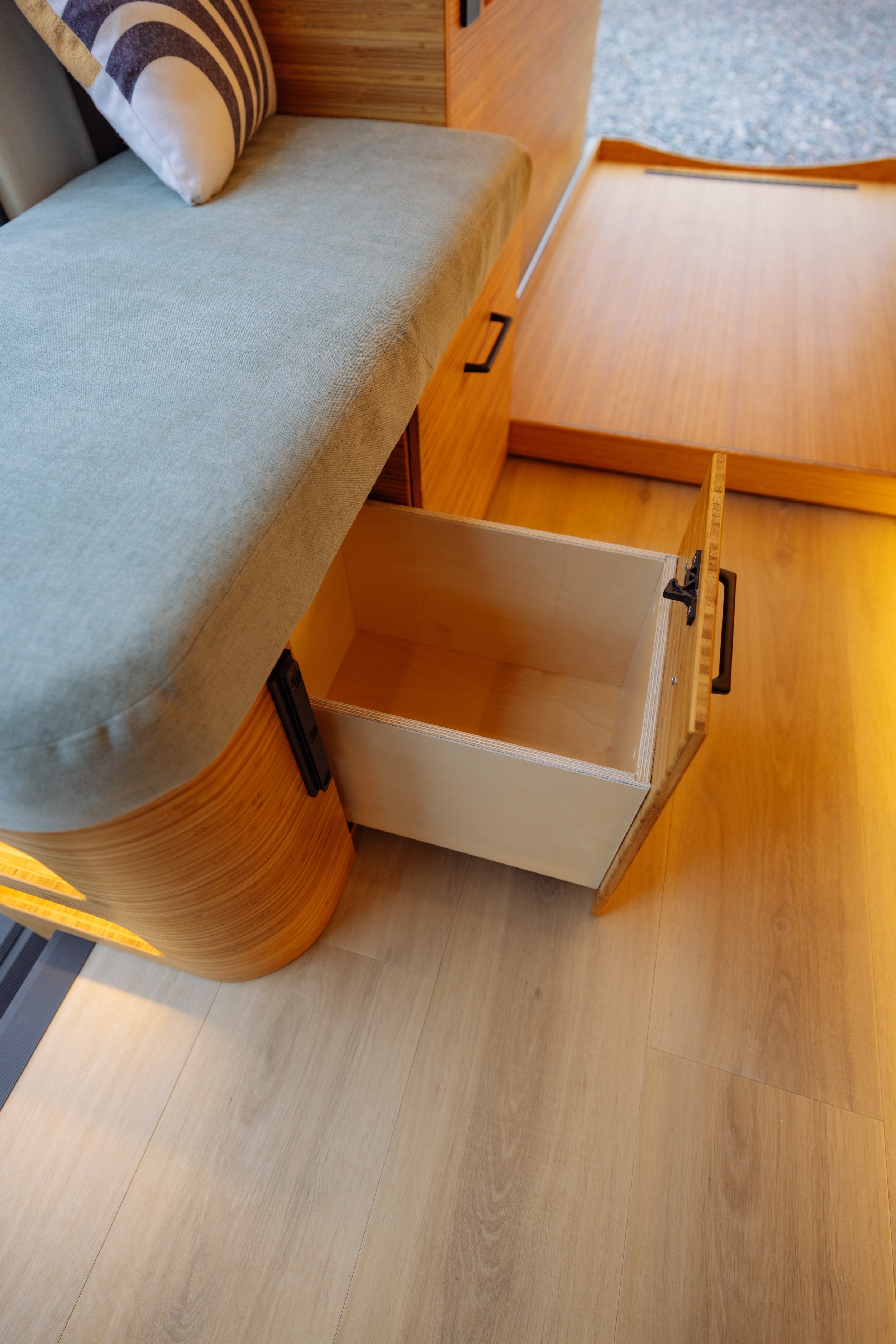Interior of a camper van with an open wooden storage drawer under a cushioned bench, placed on a light wooden floor.