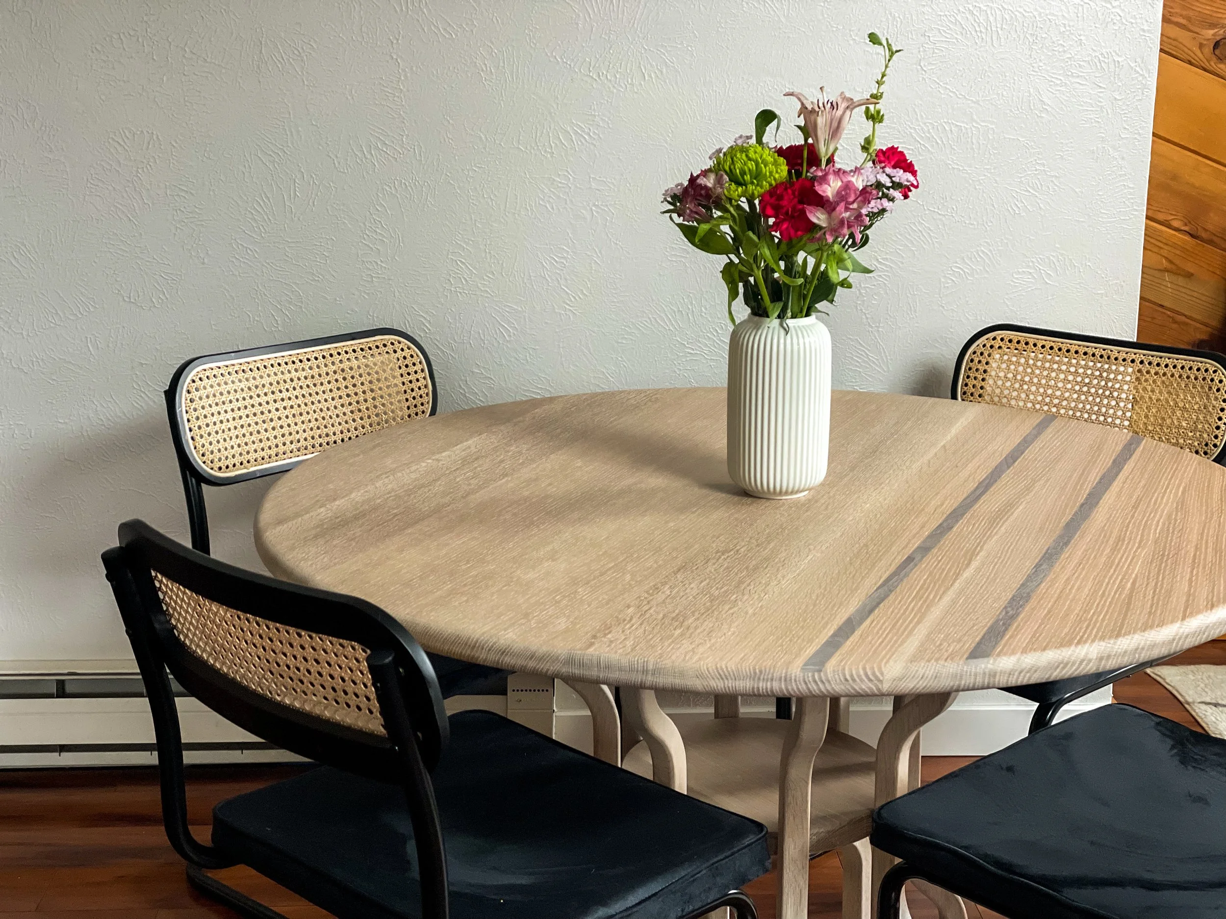 Round wooden dining table with a striped design, surrounded by chairs with cane backs and black seats. A white vase with colorful flowers is placed at the center of the table.