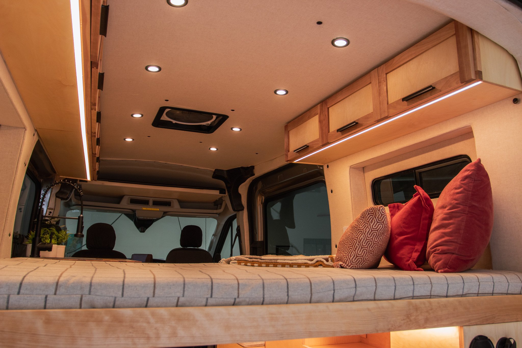 Interior of a camper van with a bed, pillows, overhead lighting, ceiling fan, and wooden storage cabinets.