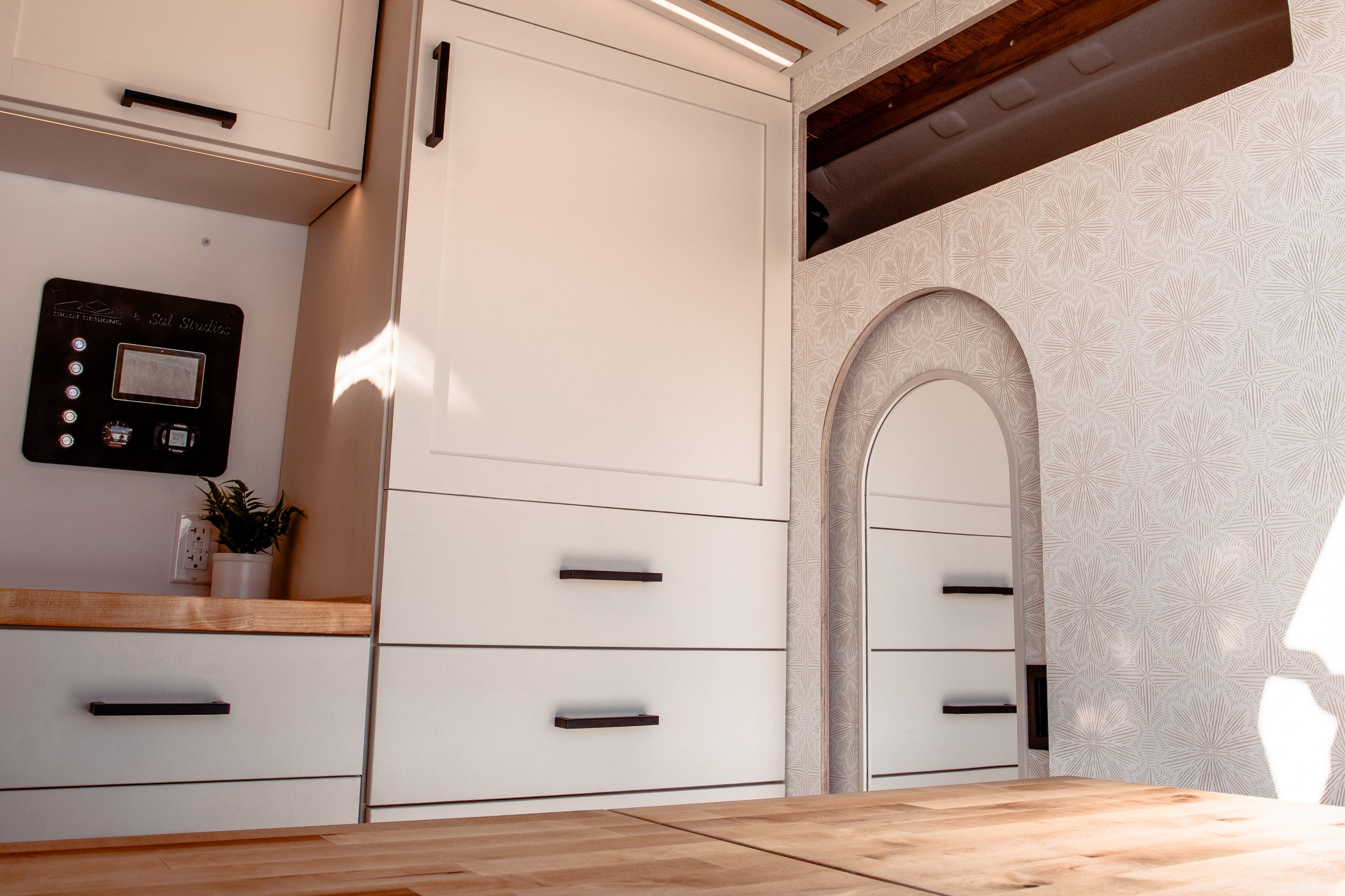 Interior of a modern camper van with white cabinets, control panel, small plant, and decorative wall paneling with a mirror and a pocket door.