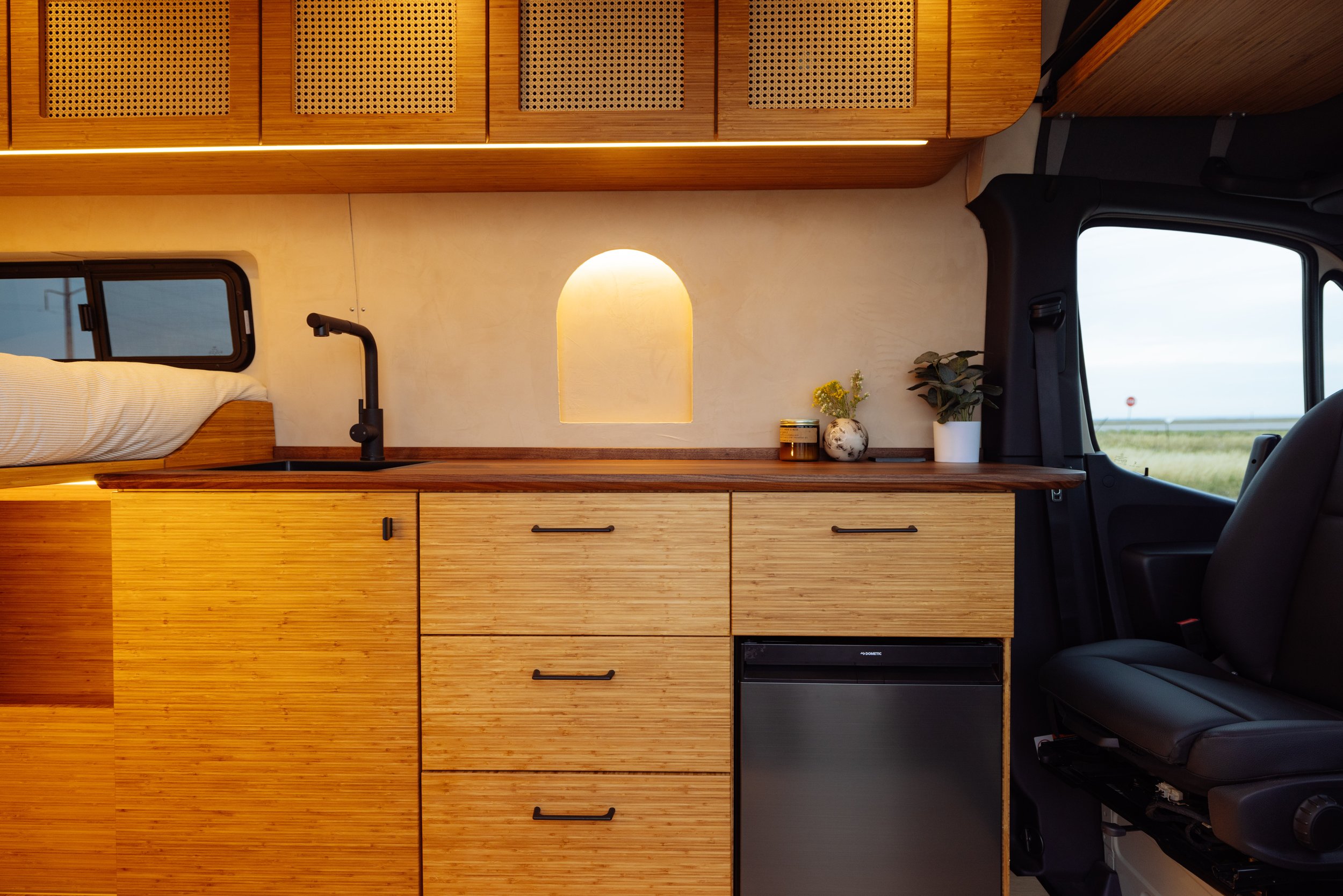 Interior of a modern camper van with stylish wooden cabinets, drawers, a small sink, and a mini refrigerator. Decorative items are placed on the countertop, creating a cozy and functional living space.