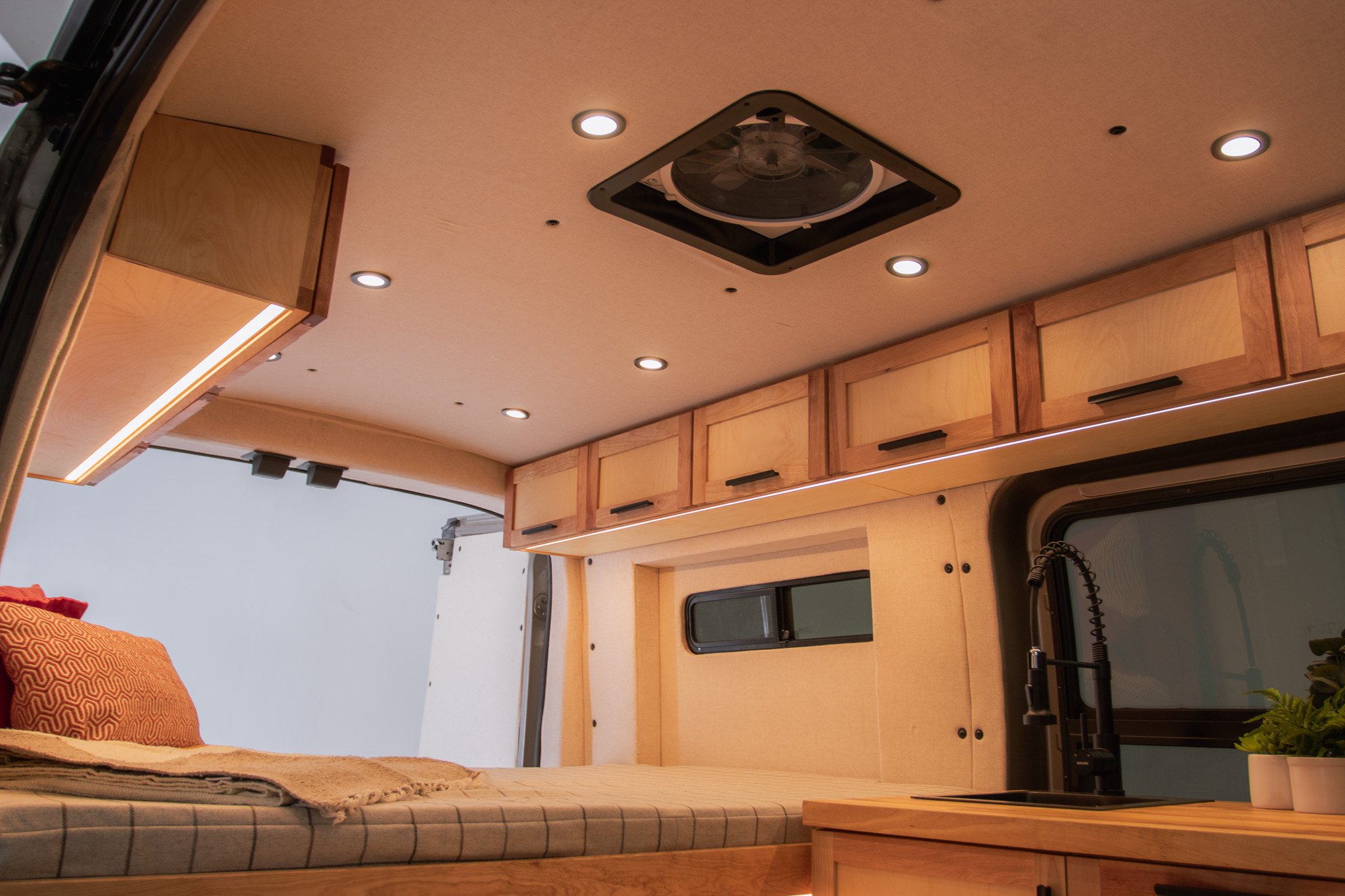 Interior of a camper van with wooden cabinets, recessed lighting, a bed with pillows and blankets, and a small kitchen area with a sink and faucet.