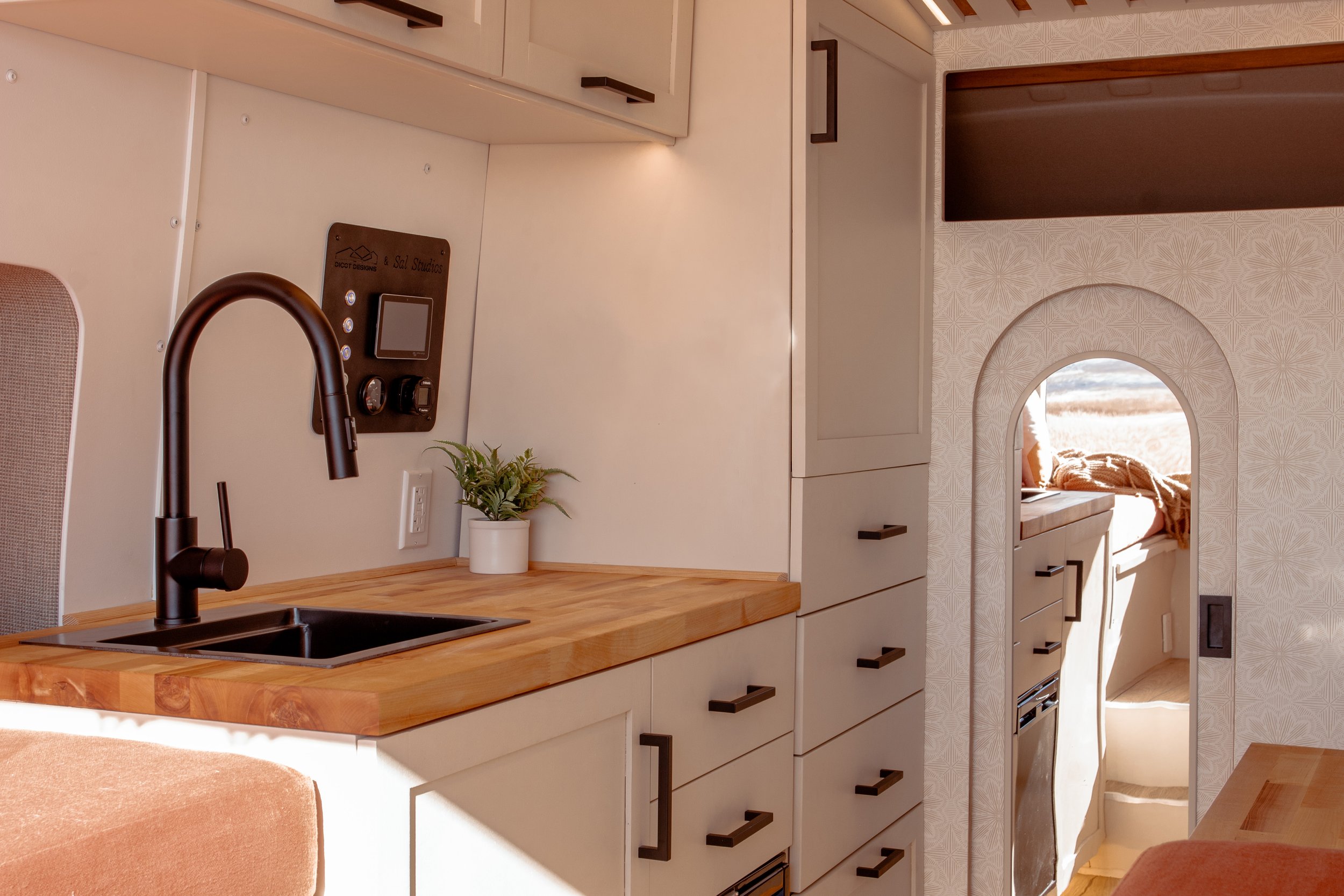 Interior of the Joy camper van featuring a compact kitchen with wooden countertops, a black faucet, and built-in cabinetry. A small potted plant sits on the counter. There's an arched doorway leading to the living area with outside views.