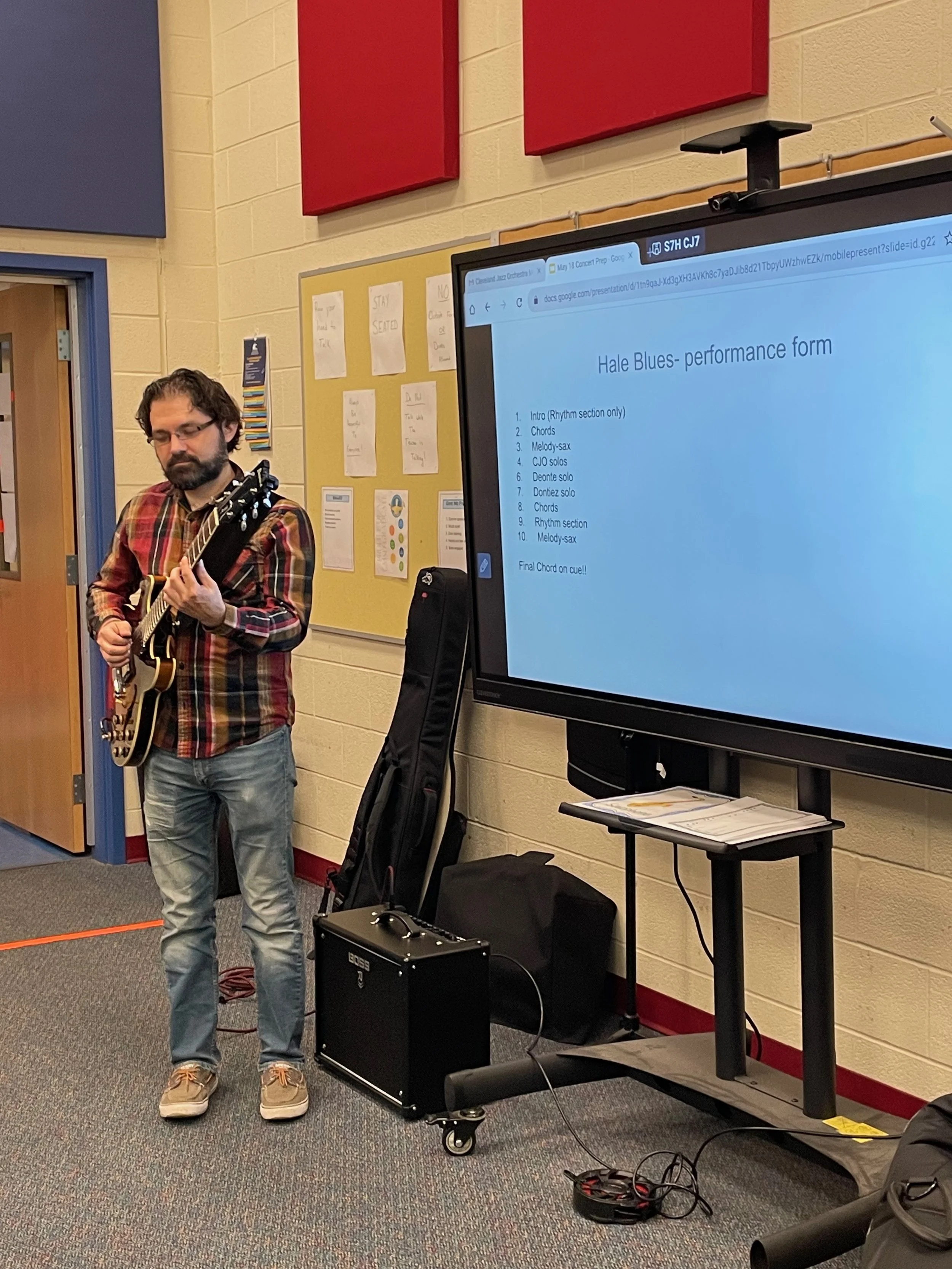 A man playing an electric guitar during a performance in a classroom. A large screen displays a playlist for a song titled 'Hale Blues,' with sections for chords and solos. The room has colorful walls, a bulletin board with papers, and equipment including a guitar case, amplifier, and a black bag.