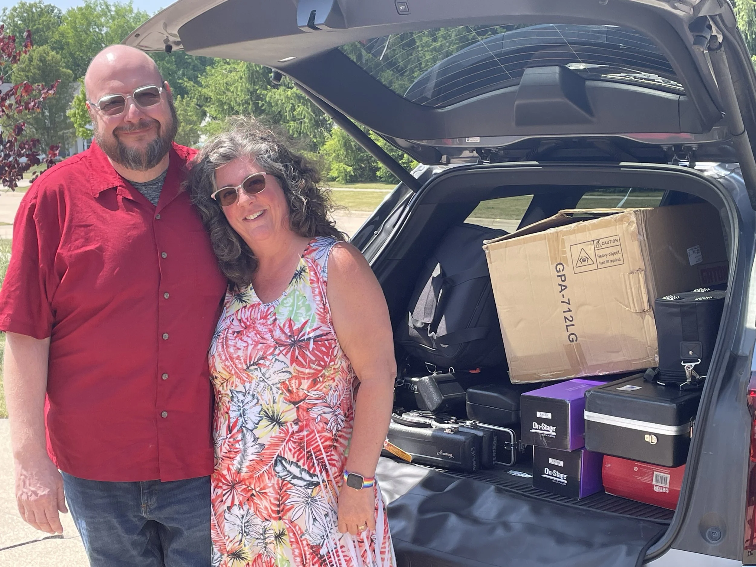 A man and woman standing outdoors with their car loaded with moving boxes and supplies, smiling at the camera.