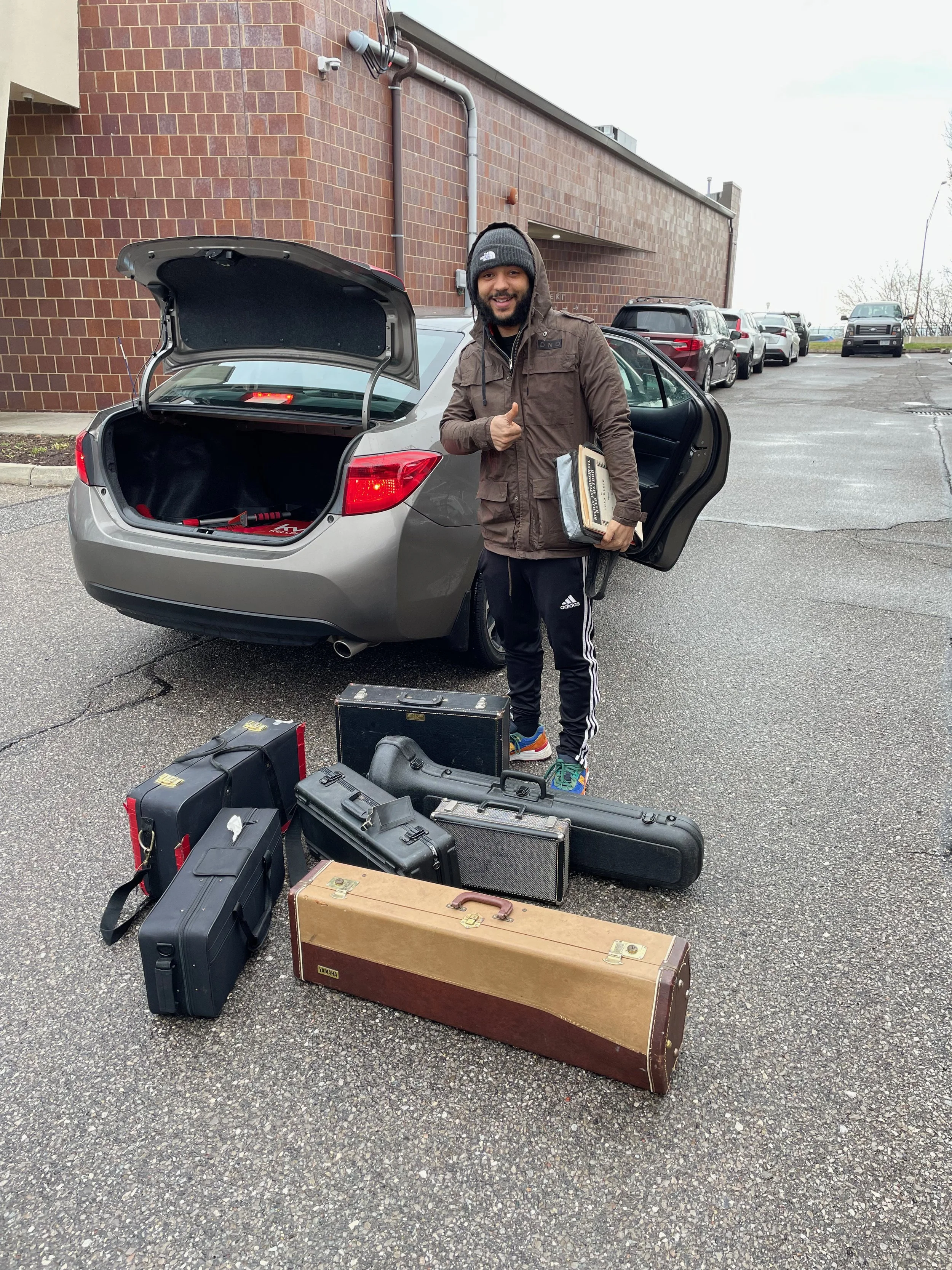A man standing next to an open car trunk, giving a thumbs up, with musical instrument cases on the ground in front of him.