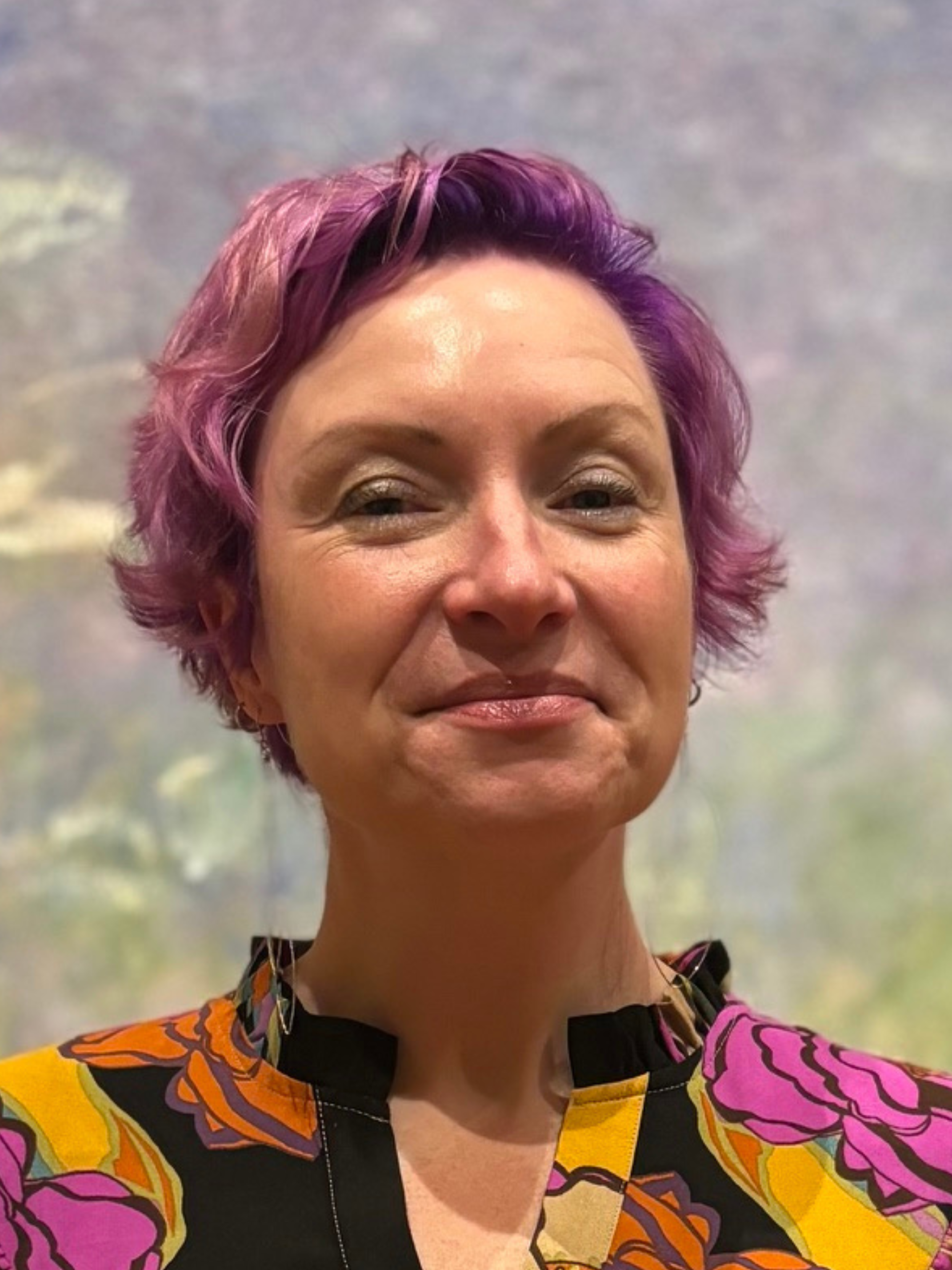 A woman with short curly purple hair smiling, wearing a colorful blouse