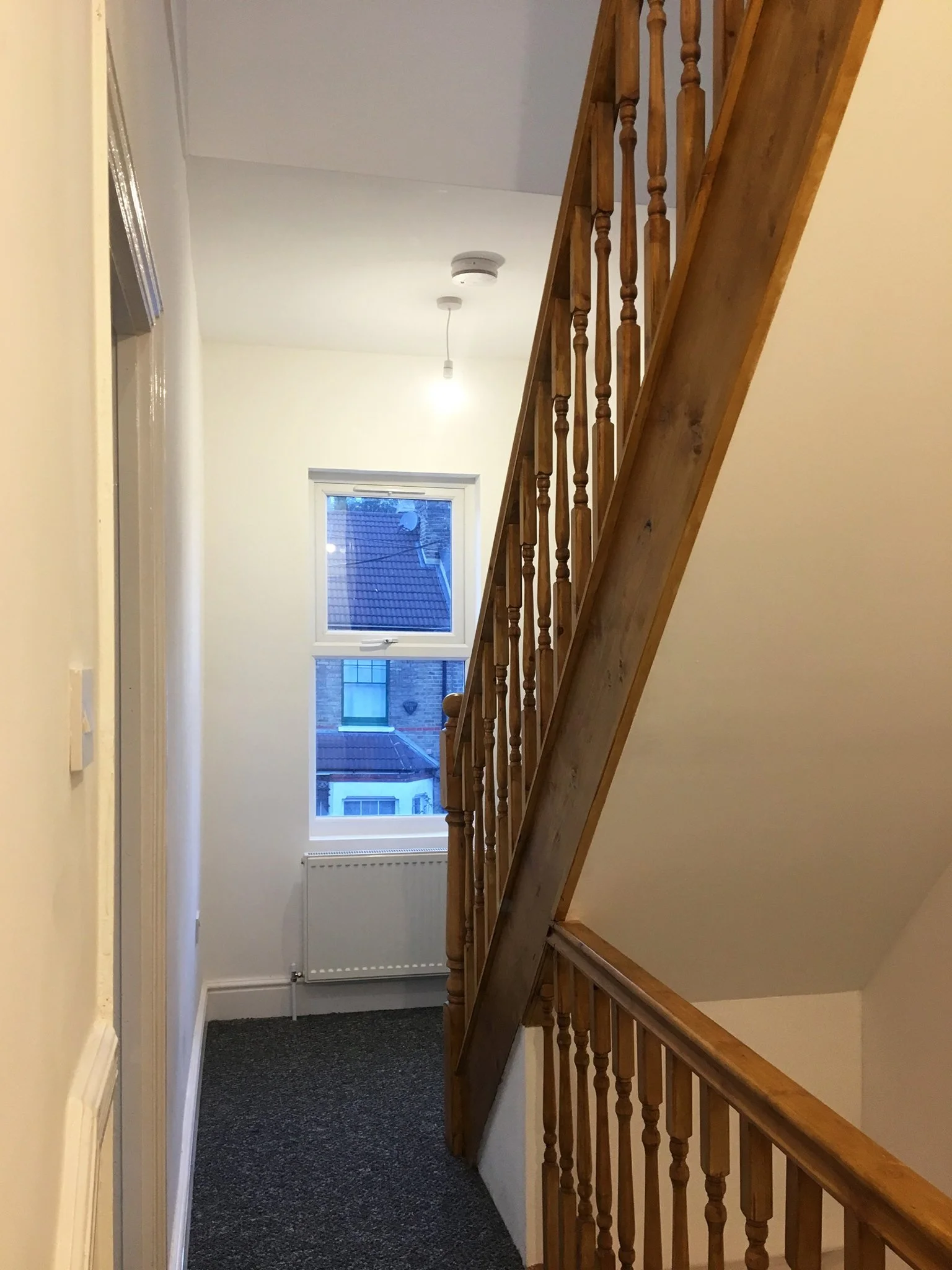 A staircase area illuminated by natural light from a window. It features wooden railings, white walls, and a view of neighbouring rooftops through the hallway window.