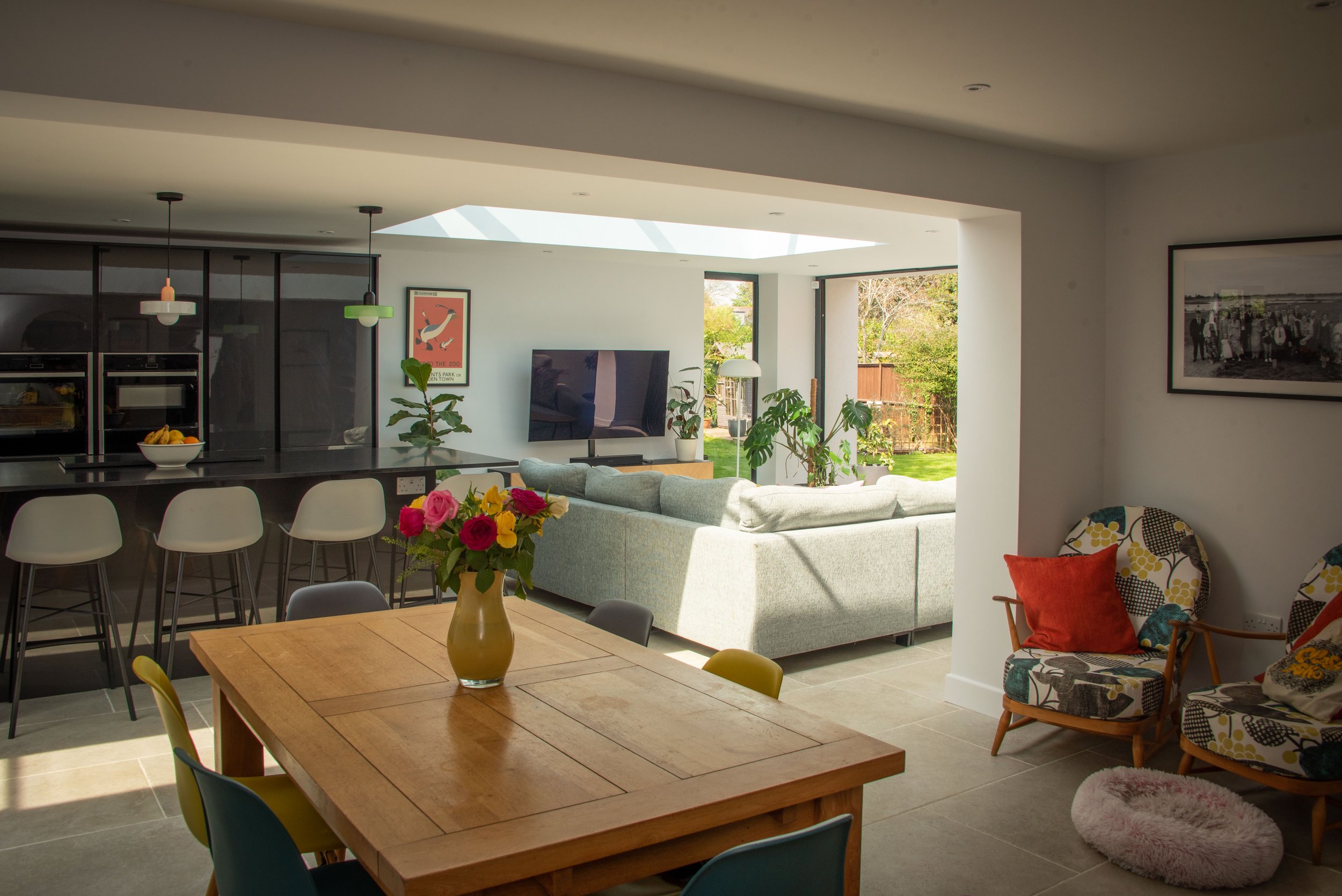 The image features a modern open-plan living and dining area. A wooden dining table with colourful chairs and a vase of flowers is in the foreground. In the background, there's a kitchen island with barstools, a grey sectional sofa, a wall-mounted TV