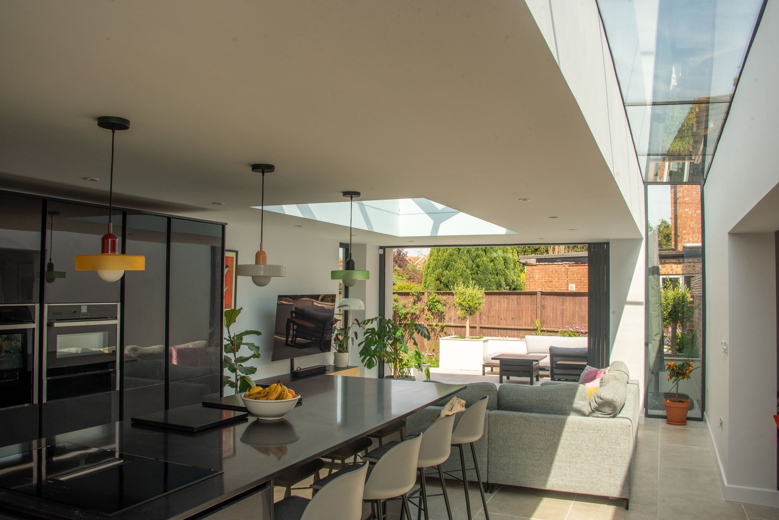 A modern, open-plan living space with a large skylight. The foreground features a kitchen island with barstools and a bowl of bananas. The living area has a grey sectional sofa with colourful cushions and is decorated with several potted plants. Floo