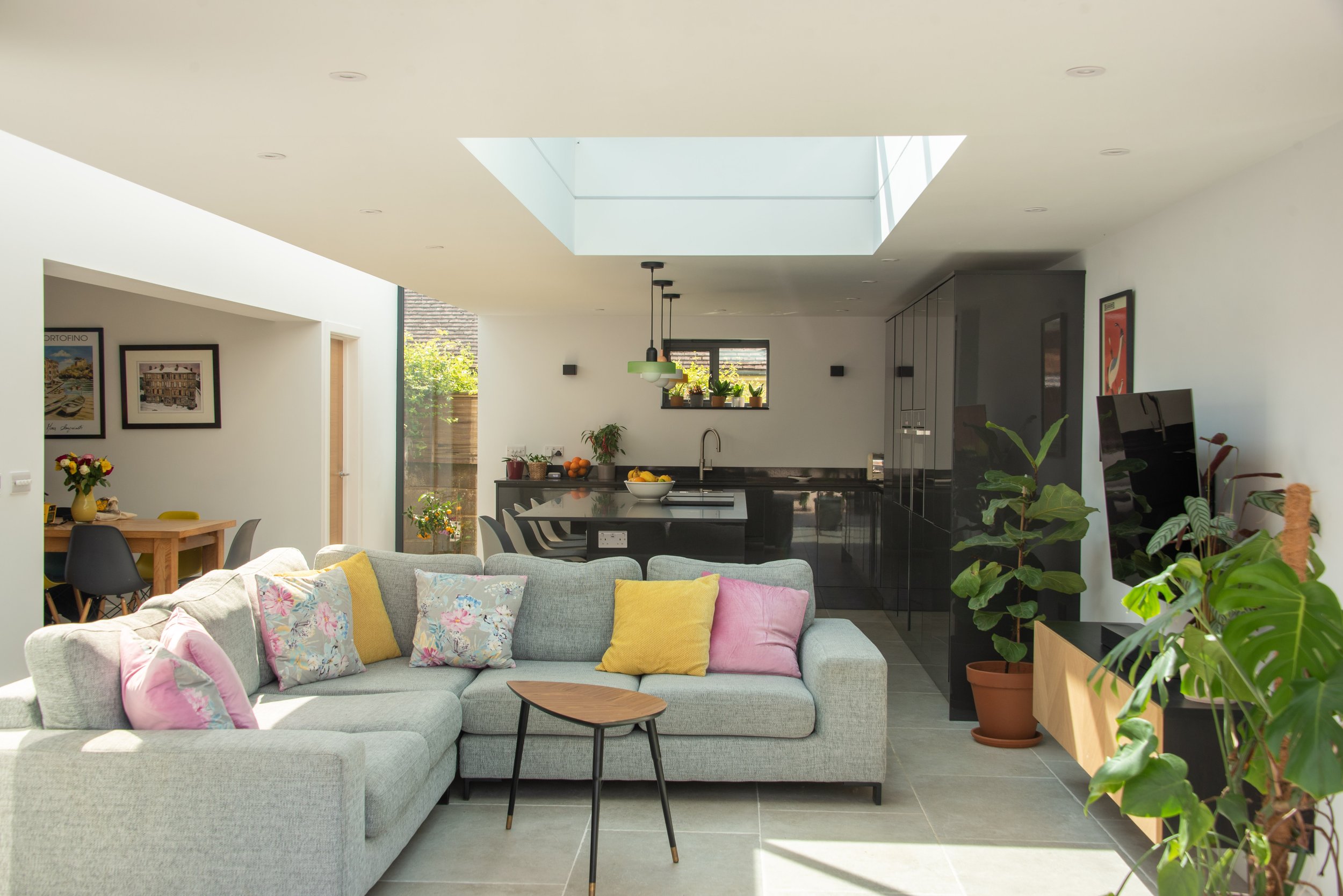 A spacious, contemporary living area featuring a large grey sectional sofa adorned with colourful throw pillows and a small wooden coffee table. The space includes various potted plants and leads into a sleek kitchen with a black countertop and cabin