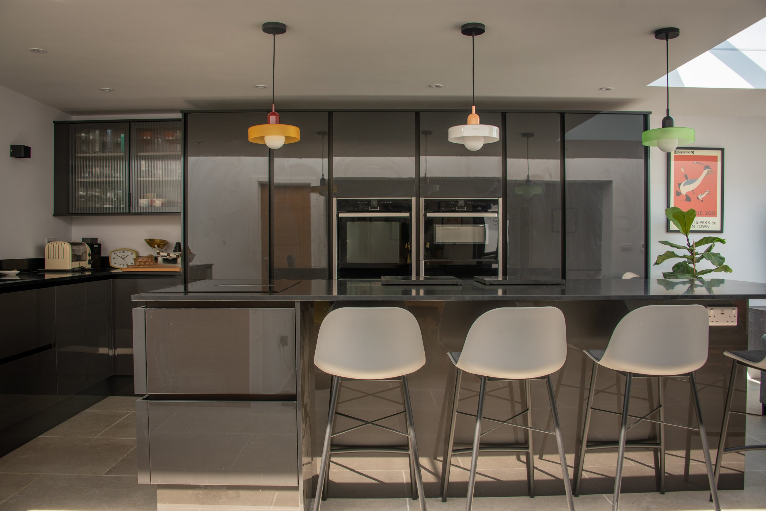 A modern, open-plan living space with a large skylight. The foreground features a kitchen island with barstools. Three lights hang above the kitchen island with yellow, white, and green lampshades. In the background, there are floor-to-ceiling cabine
