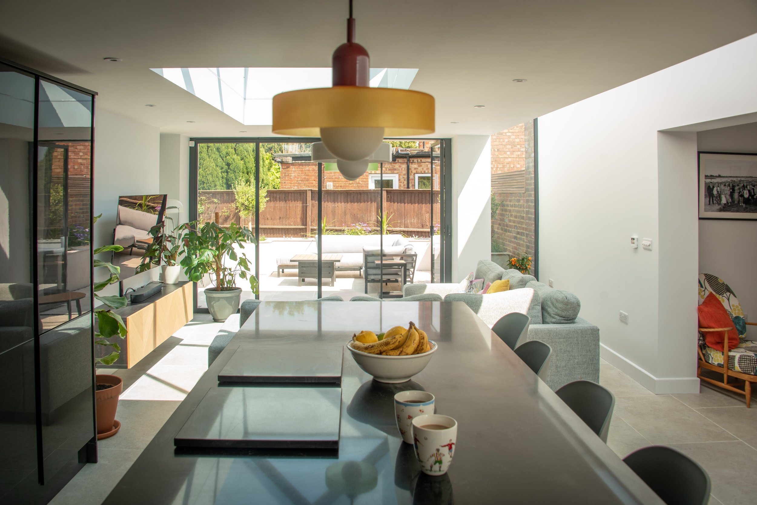 A modern, open-plan living space with a large skylight. The foreground features a kitchen island with barstools, a bowl of bananas, and three mugs on the counter. The living area has a grey sectional sofa with colourful cushions and is decorated with