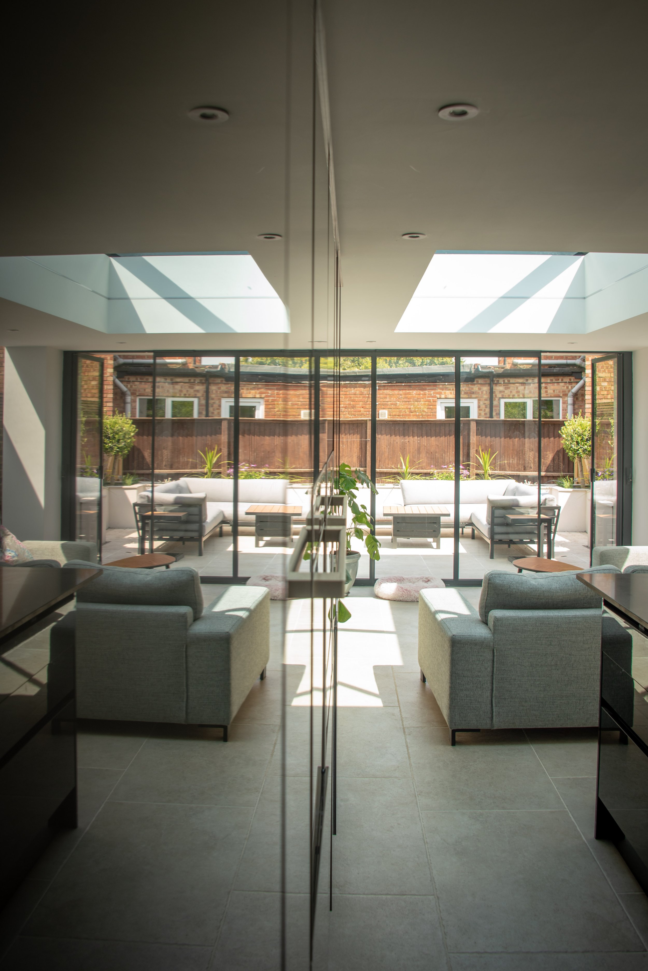 A modern living area reflected in a cupboard door showing the corner of a grey sectional sofa facing a floor-to-ceiling glass doors. The space is illuminated by two large skylights, allowing natural light to flood the room. Beyond the glass wall, the
