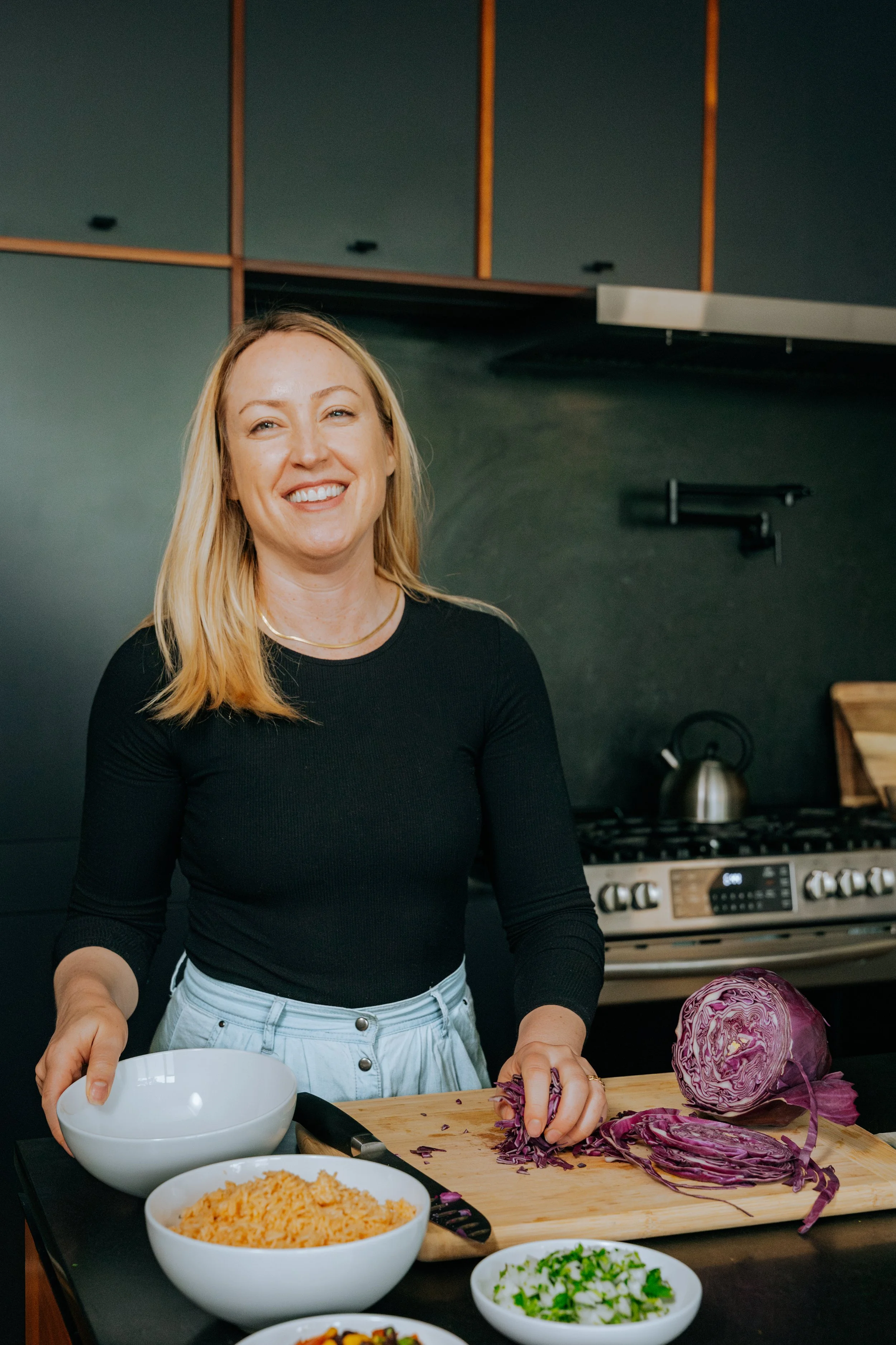 Image of a white, blond woman with blue eyes in a long-sleeve black top and denim bottoms smiling and preparing chopped purple cabbage in a dark kitchen, preparing organic meals in Chicago