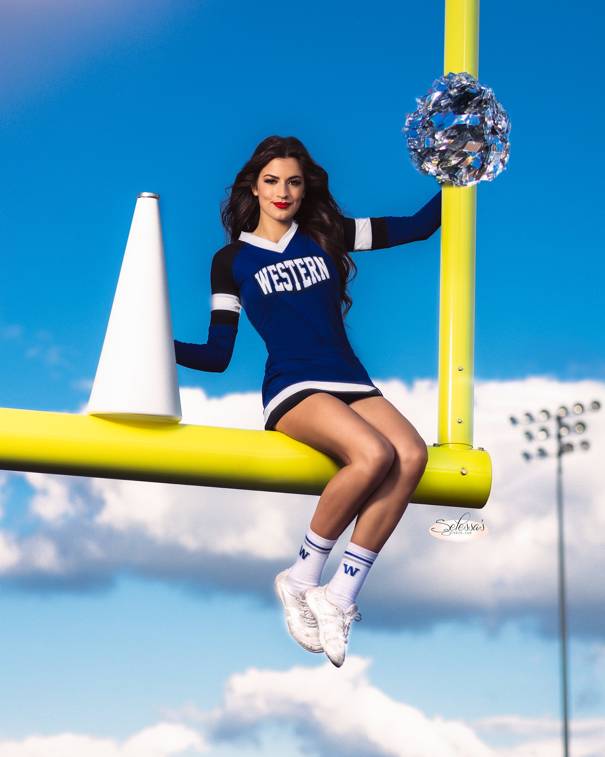 Cheerleader in blue and white uniform sitting on a yellow goalpost, holding pom-poms, with a megaphone nearby. Sky and clouds in the background.
