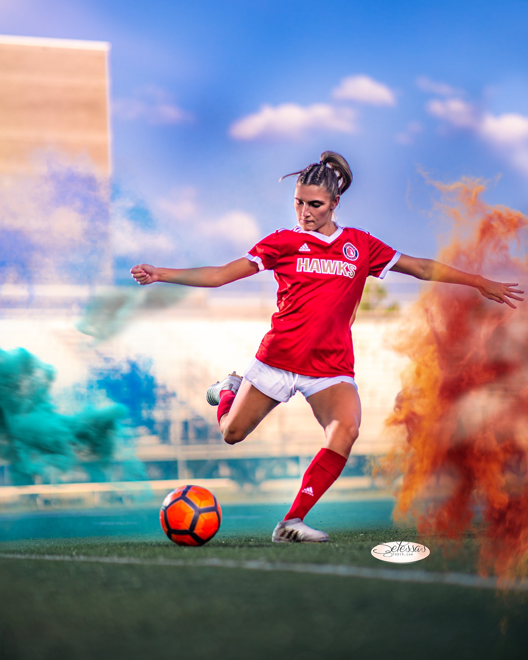 A female soccer player in red uniform kicking a soccer ball on a field, surrounded by colorful smoke.