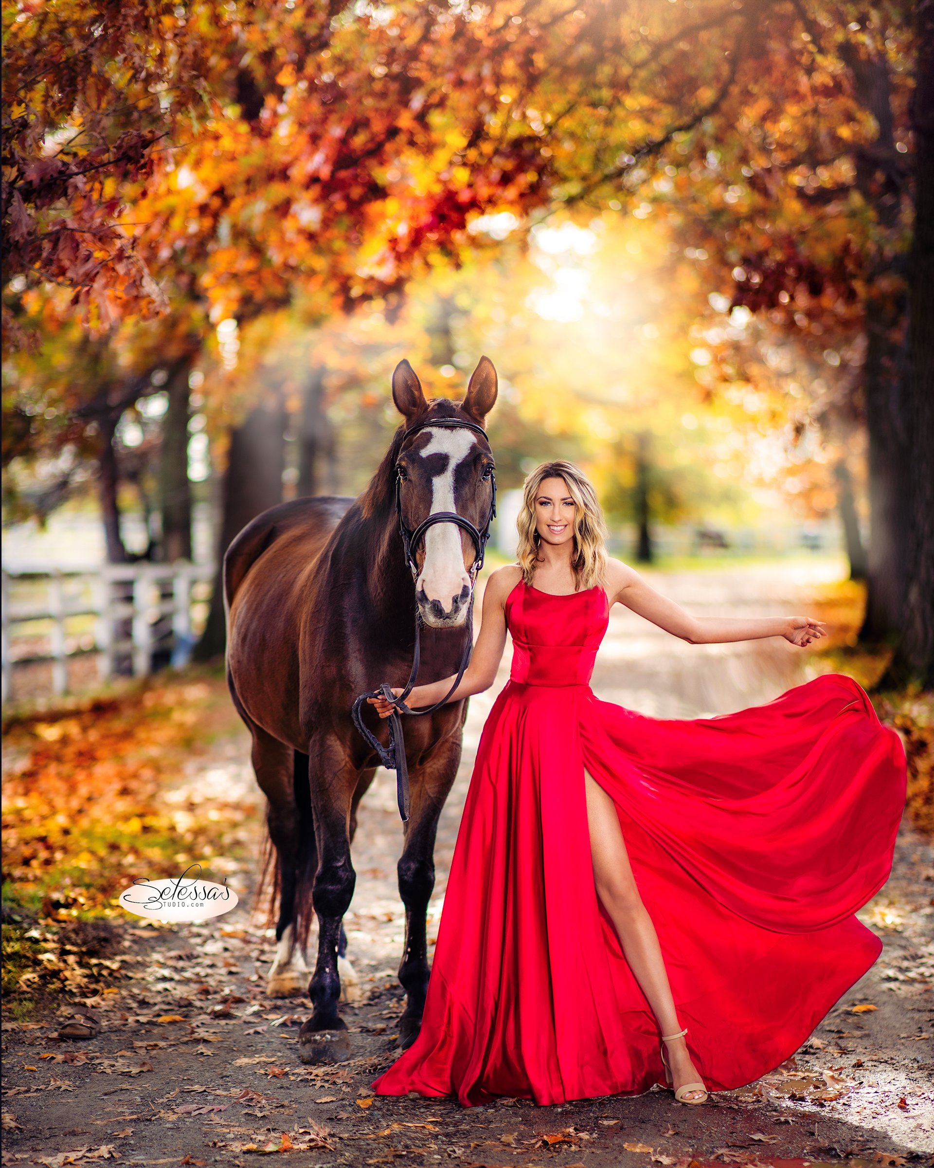 graduating senior girl in a flowing red dress standing beside a horse on a leafy path during autumn.