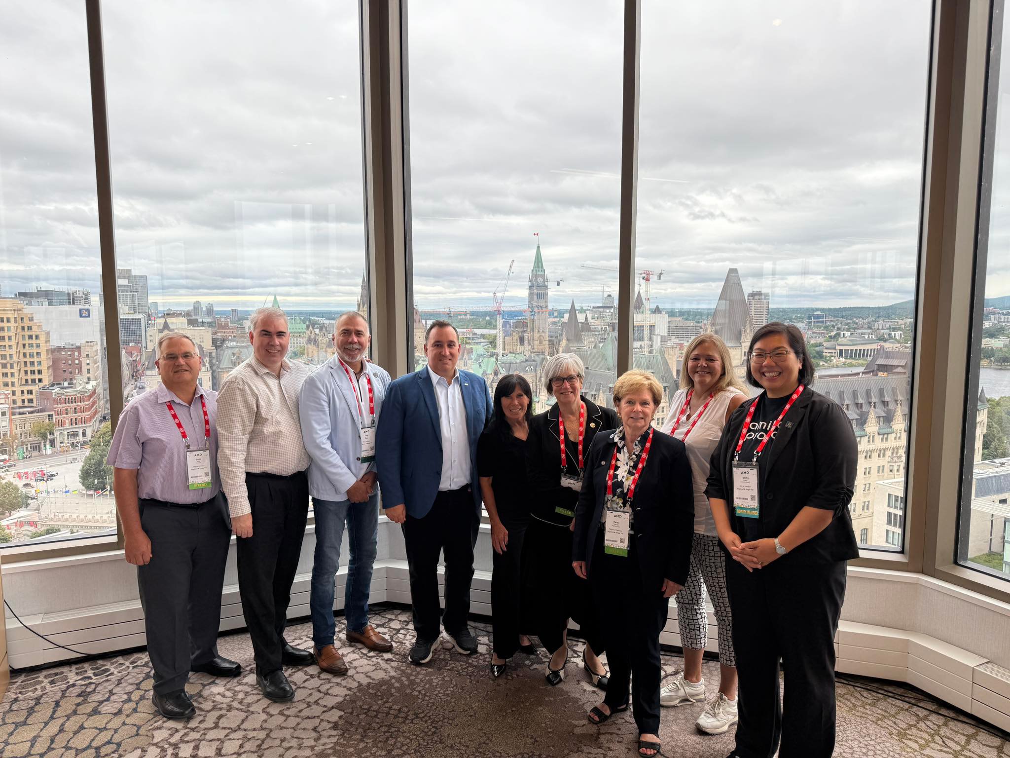 Group of ten people standing in front of a large window with a city skyline view, including iconic buildings and cloudy sky.