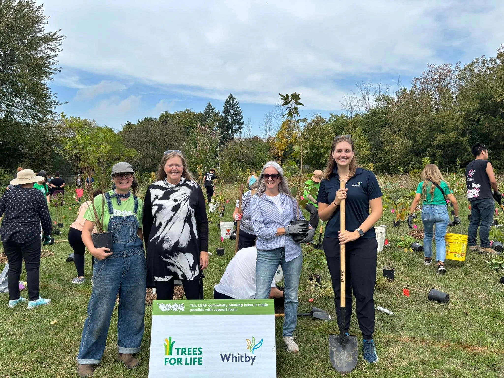 Four women standing outdoors at a community tree planting event, smiling, with other people and trees in the background. Some women are holding shovels and wearing gloves, and a sign in front promotes trees for life with support from Whitby.