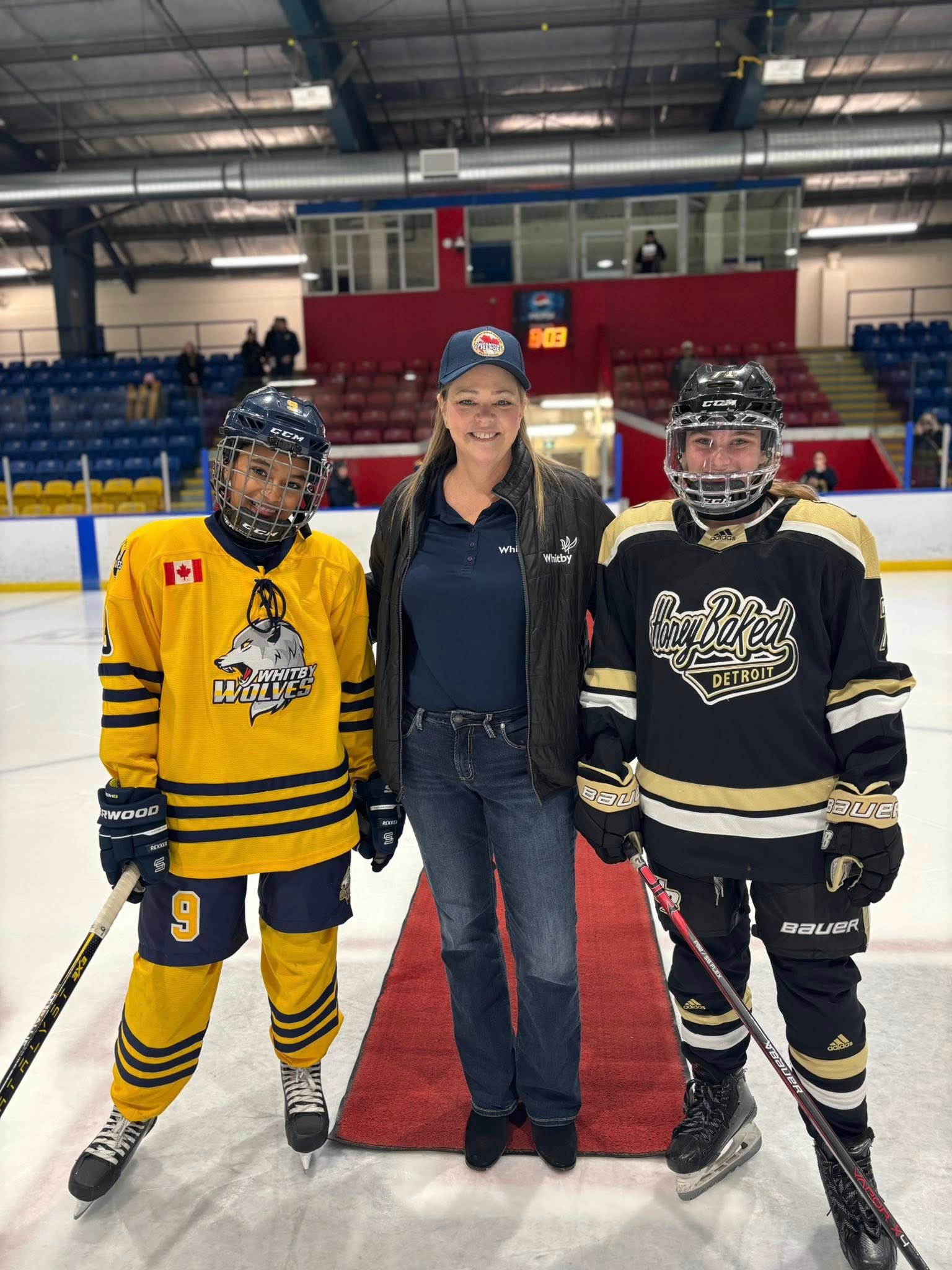 Two young hockey players in yellow and black jerseys with black helmets, standing on ice rink with a woman in the middle, all smiling for the photo. The rink has empty red and blue seats and a few spectators.