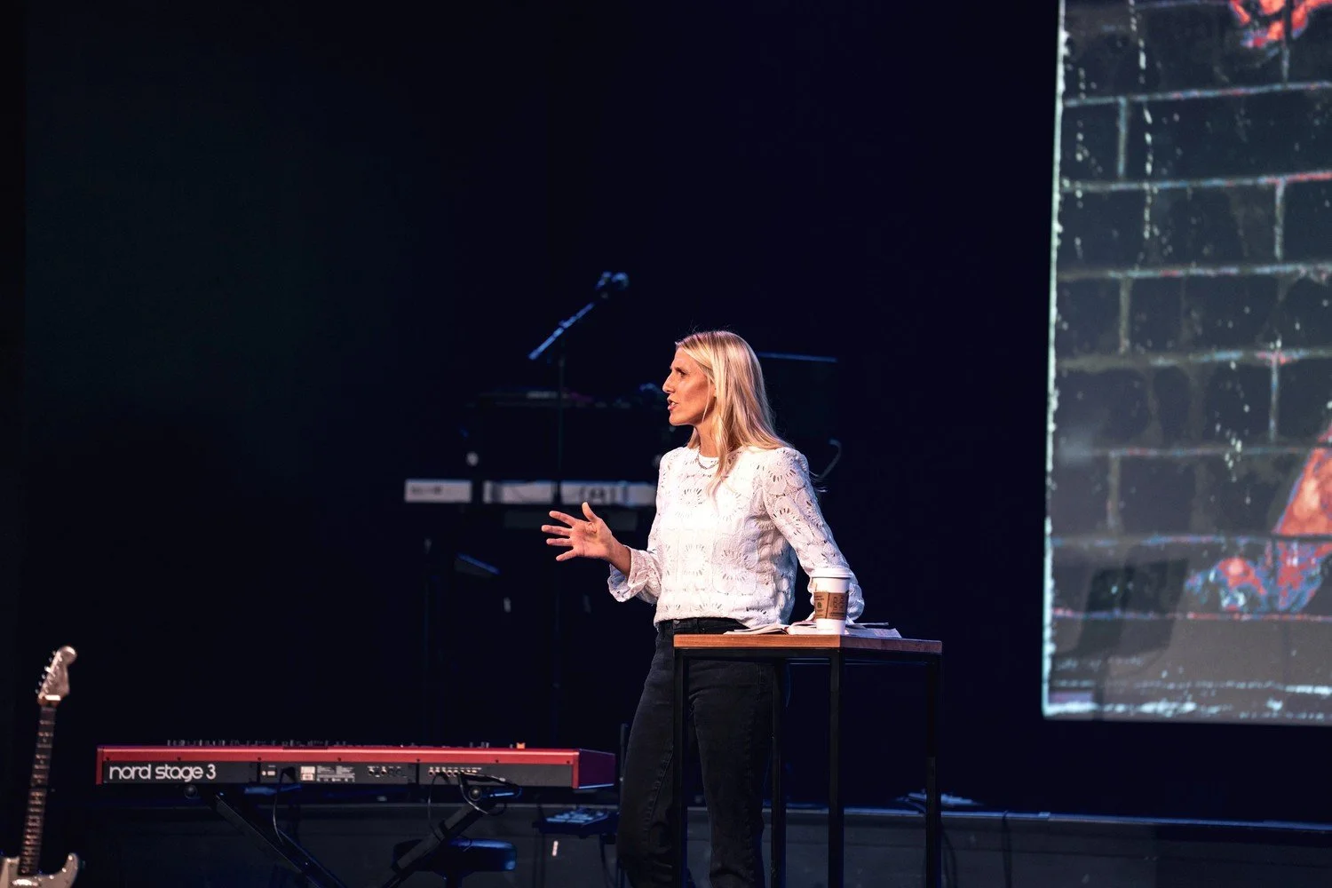 A woman with blonde hair wearing a white blouse and black pants speaking on stage during a presentation, with a keyboard and a guitar on the left and a large screen behind her.