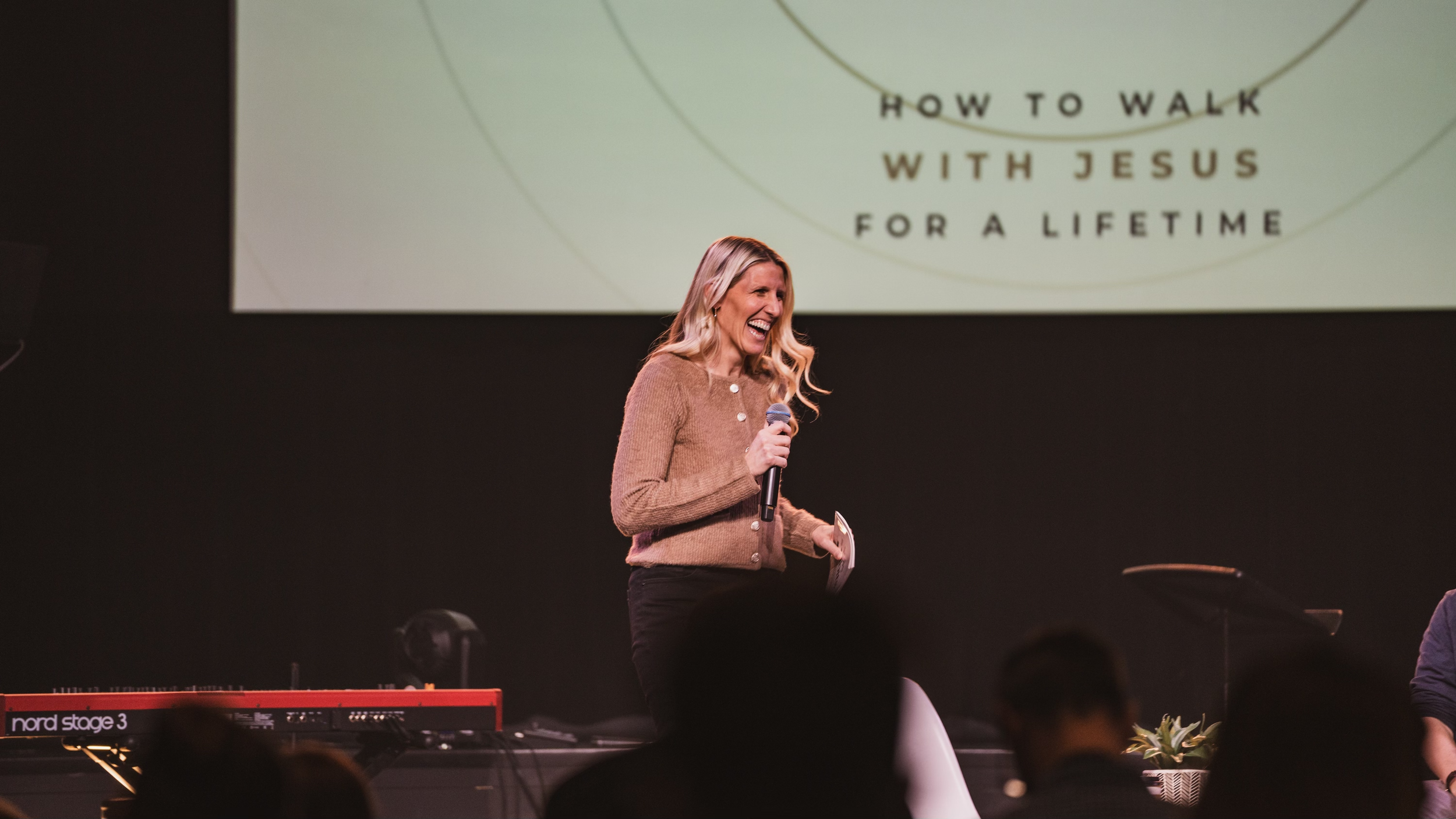 A woman with long blonde hair in a brown sweater holding a microphone and a paper, smiling on stage with a large screen behind her displaying the text 'How to Walk with Jesus for a Lifetime'.