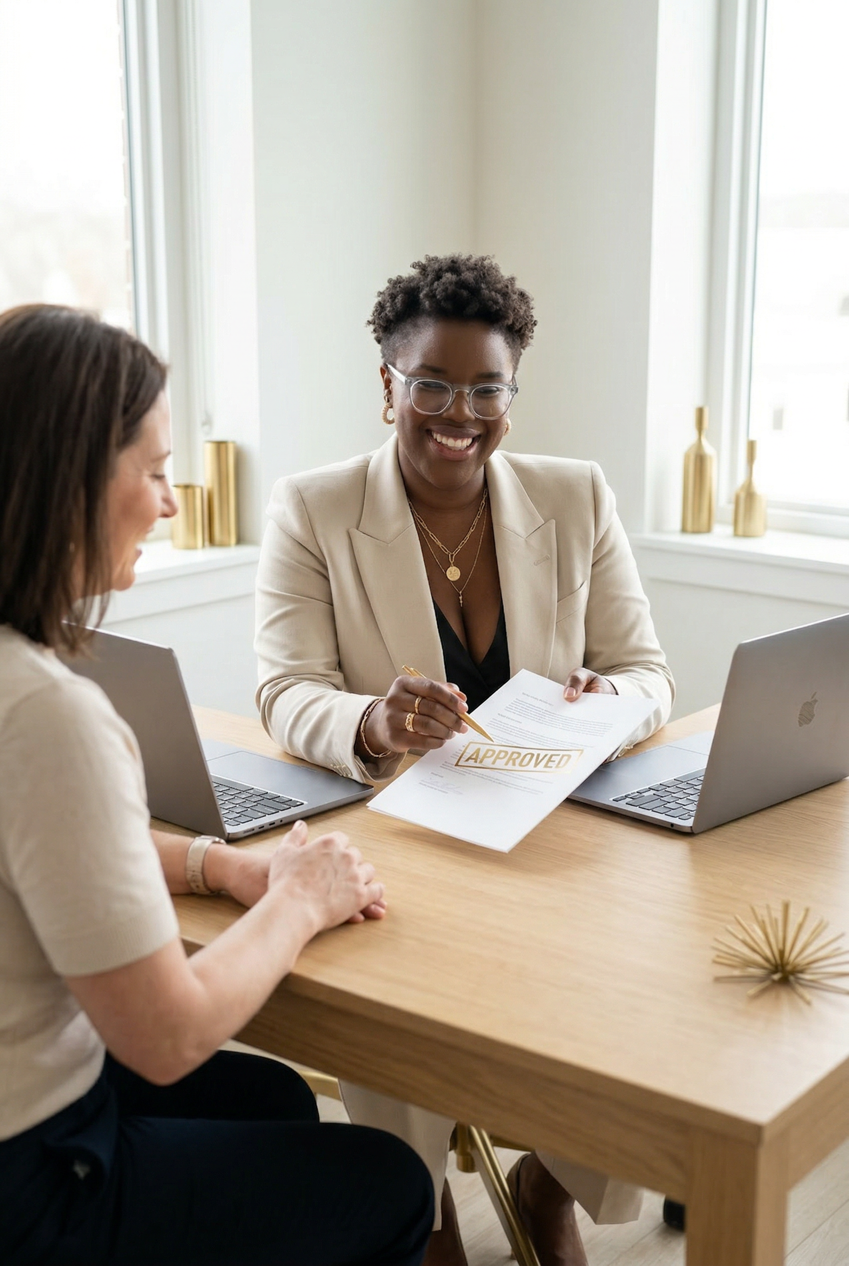 Two women in a meeting room, one is smiling and holding a document with 'APPROVED' stamped on it, while the other is listening, with laptops open in front of them.