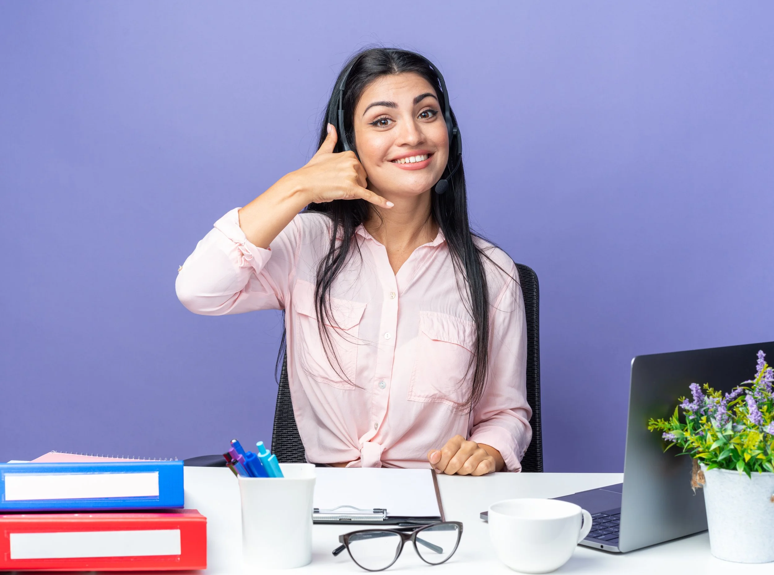young-beautiful-woman-casual-clothes-wearing-headset-with-microphone-smiling-making-call-me-gesture-sitting-table-with-laptop-blue-wall-working-office.jpg