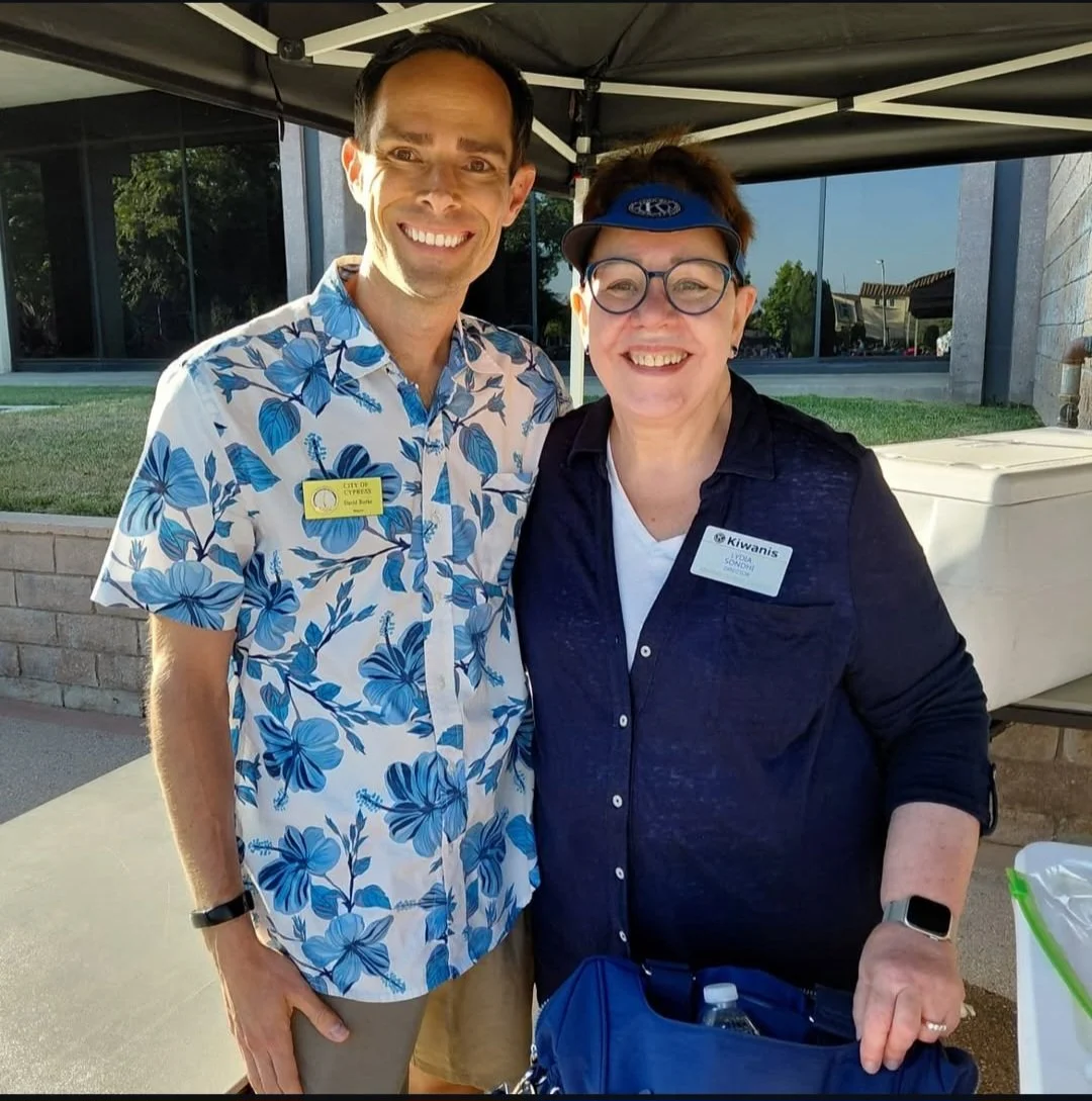 Two smiling people standing under a tent outdoors, one wearing a floral shirt and the other wearing a dark shirt with a Kiwanis badge, sunglasses, and a visor.