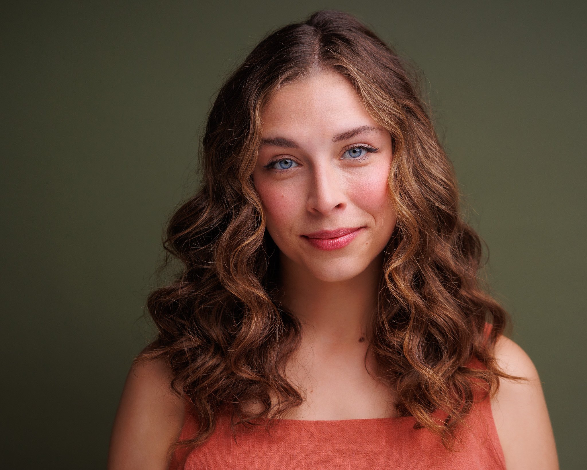 Close-up portrait of a young woman with wavy brown hair and blue eyes, wearing a sleeveless coral top, smiling softly against a plain green background.