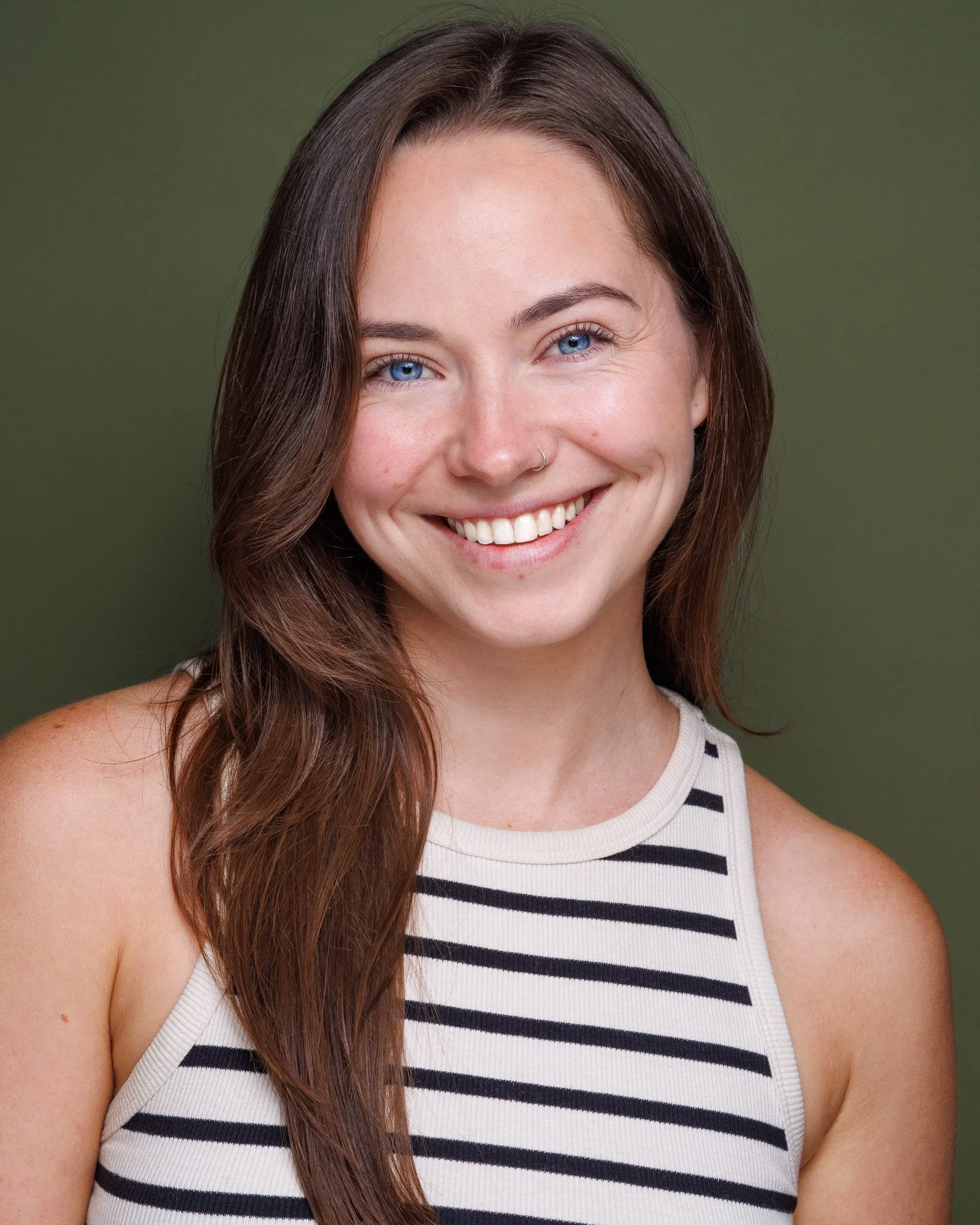 Close-up of a young woman with long brown hair, blue eyes, tan skin, wearing a white tank top with black stripes, smiling against a green background.