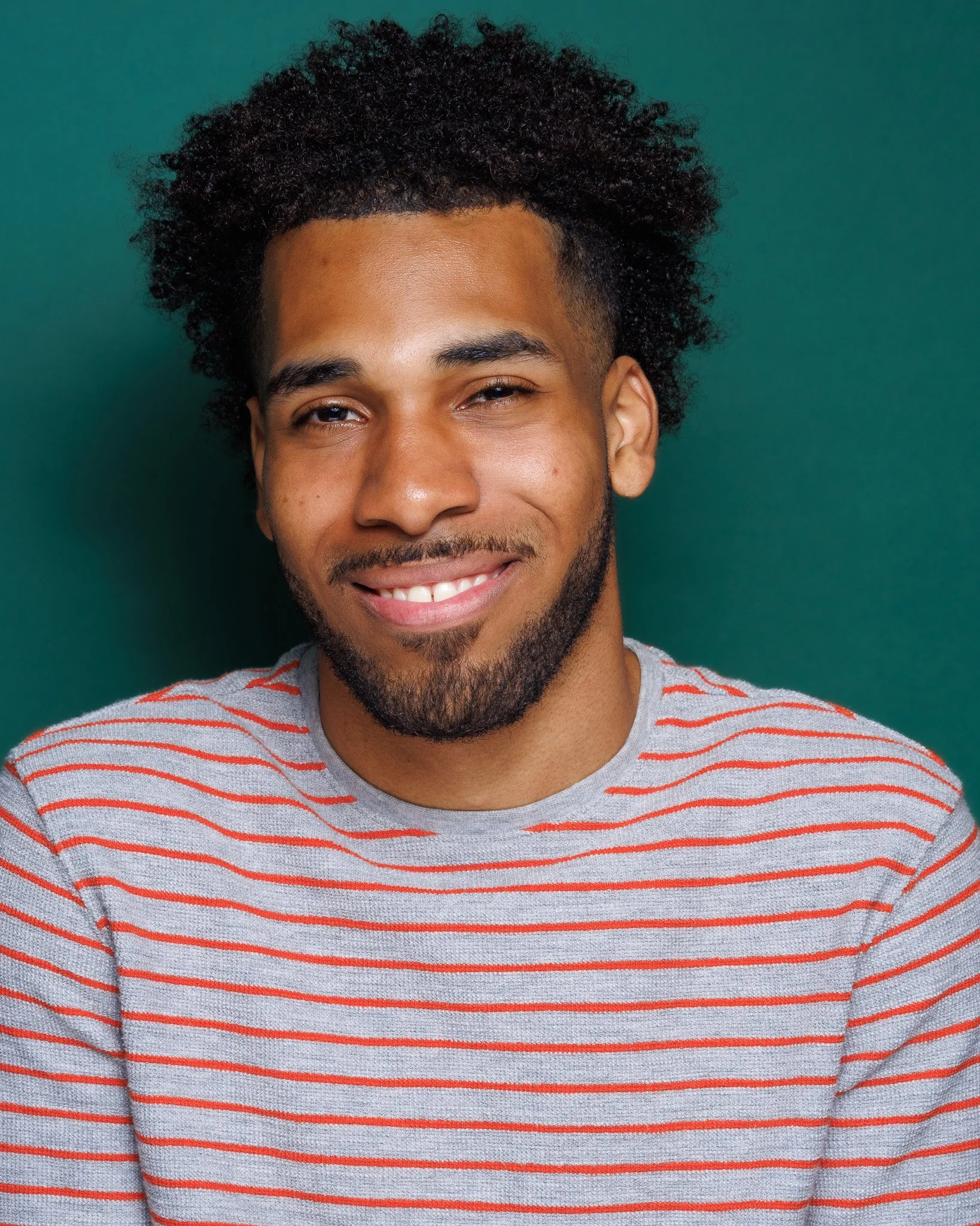 Close-up of a smiling man with curly hair and a beard, wearing a gray and red striped shirt, against a green background.