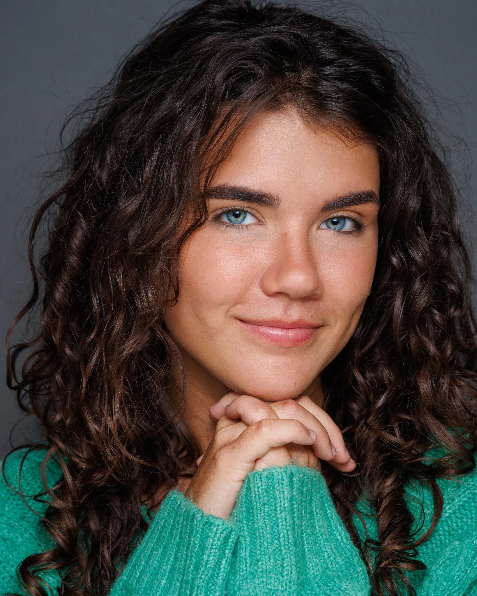 Close-up of a smiling woman with curly brown hair, wearing a green sweater, with her hands clasped under her chin.