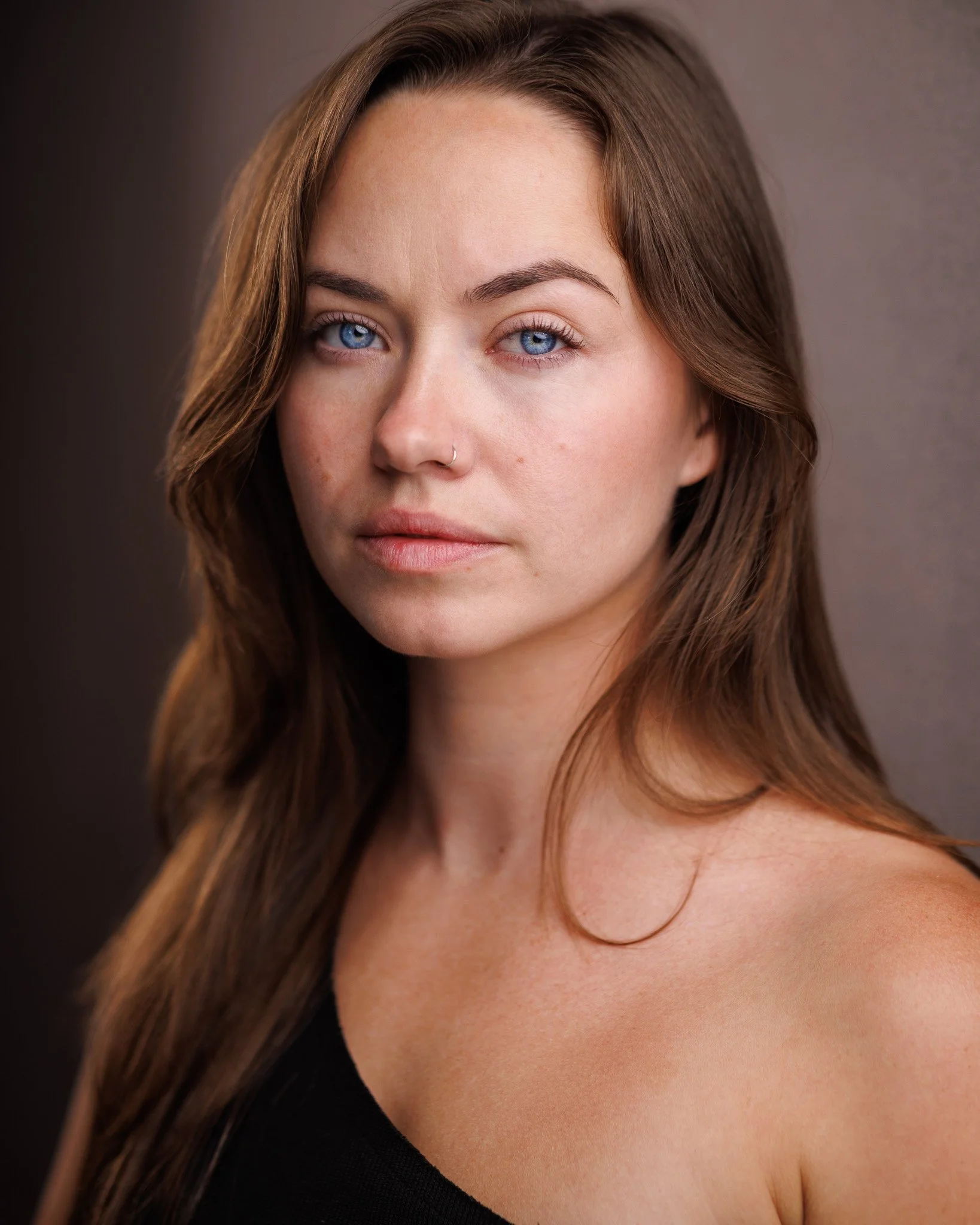 Close-up photo of a woman with blue eyes, brown hair, a small nose piercing, and natural makeup, against a dark background.