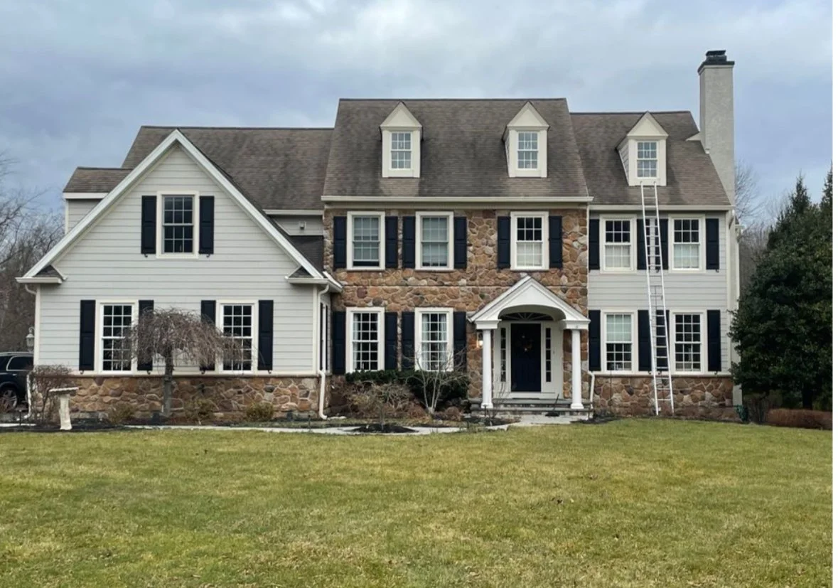 Large two-story house with a stone and white siding exterior, black shutters, multiple windows, a front doorway with columns, a ladder leaning against the right side, and a green lawn in front.